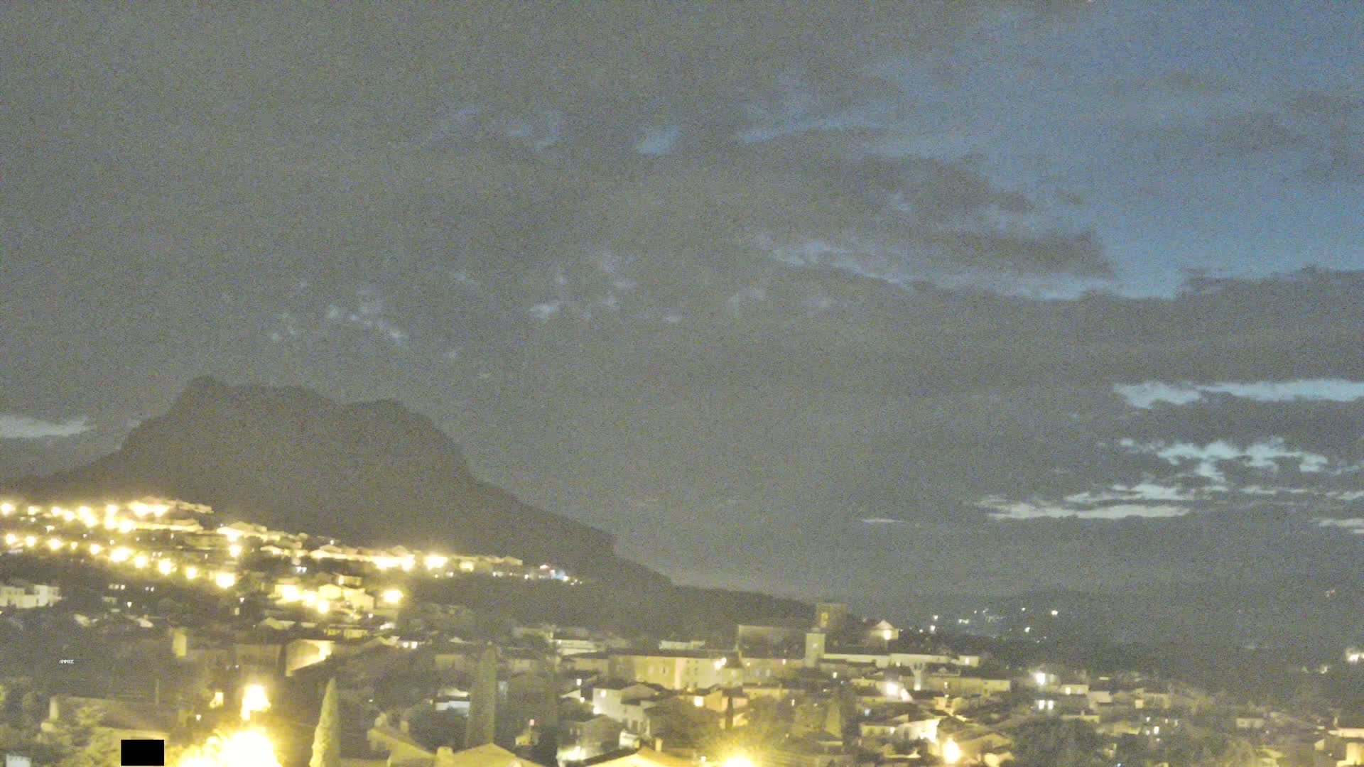 A nighttime view of a town illuminated by lights, nestled at the base of a dark mountain under a mostly cloudy sky.