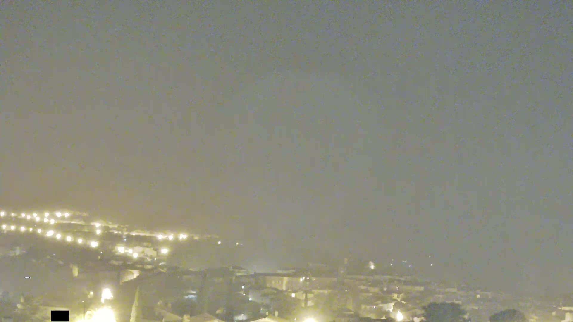 A hazy night scene shows a town's dimly lit buildings and streets from an elevated viewpoint.