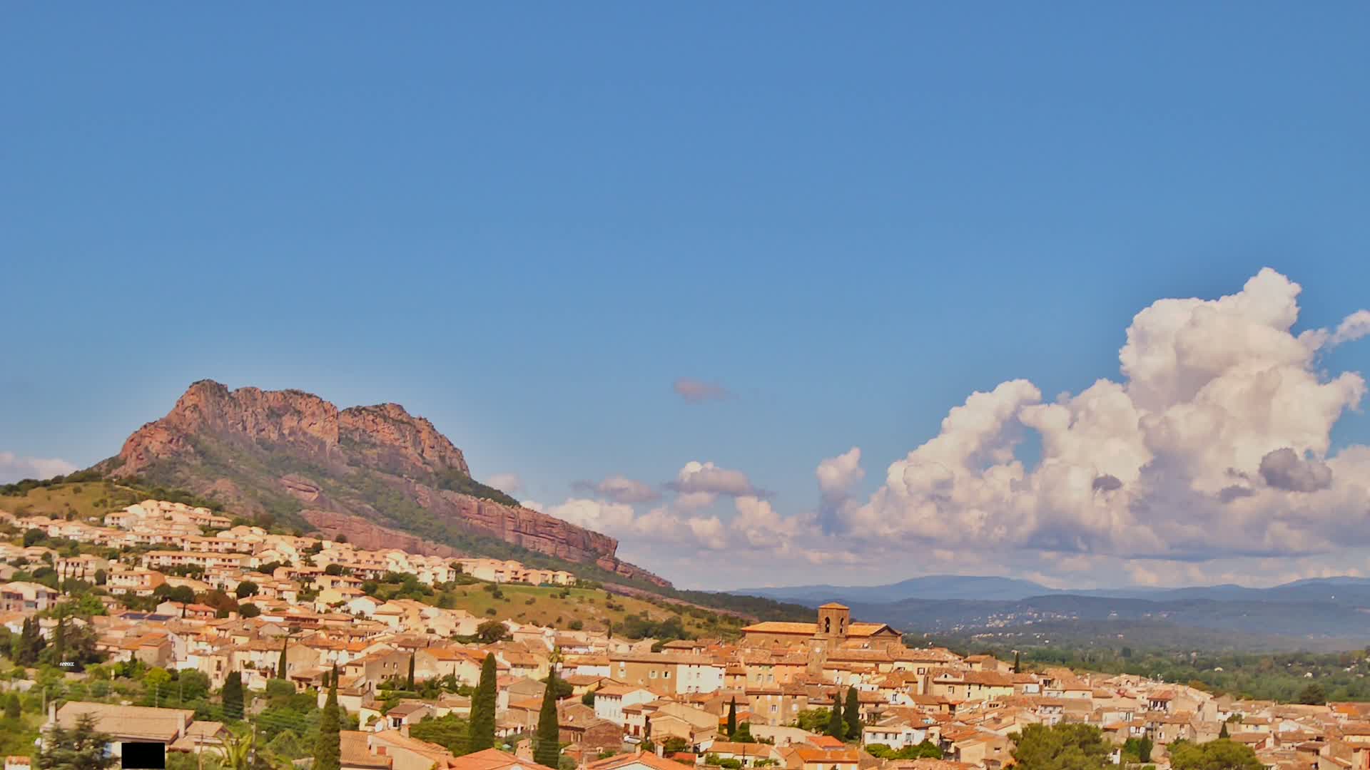 A small town with terracotta rooftops nestles at the base of a rocky, reddish-brown mountain under a mostly clear blue sky with large cumulus clouds.