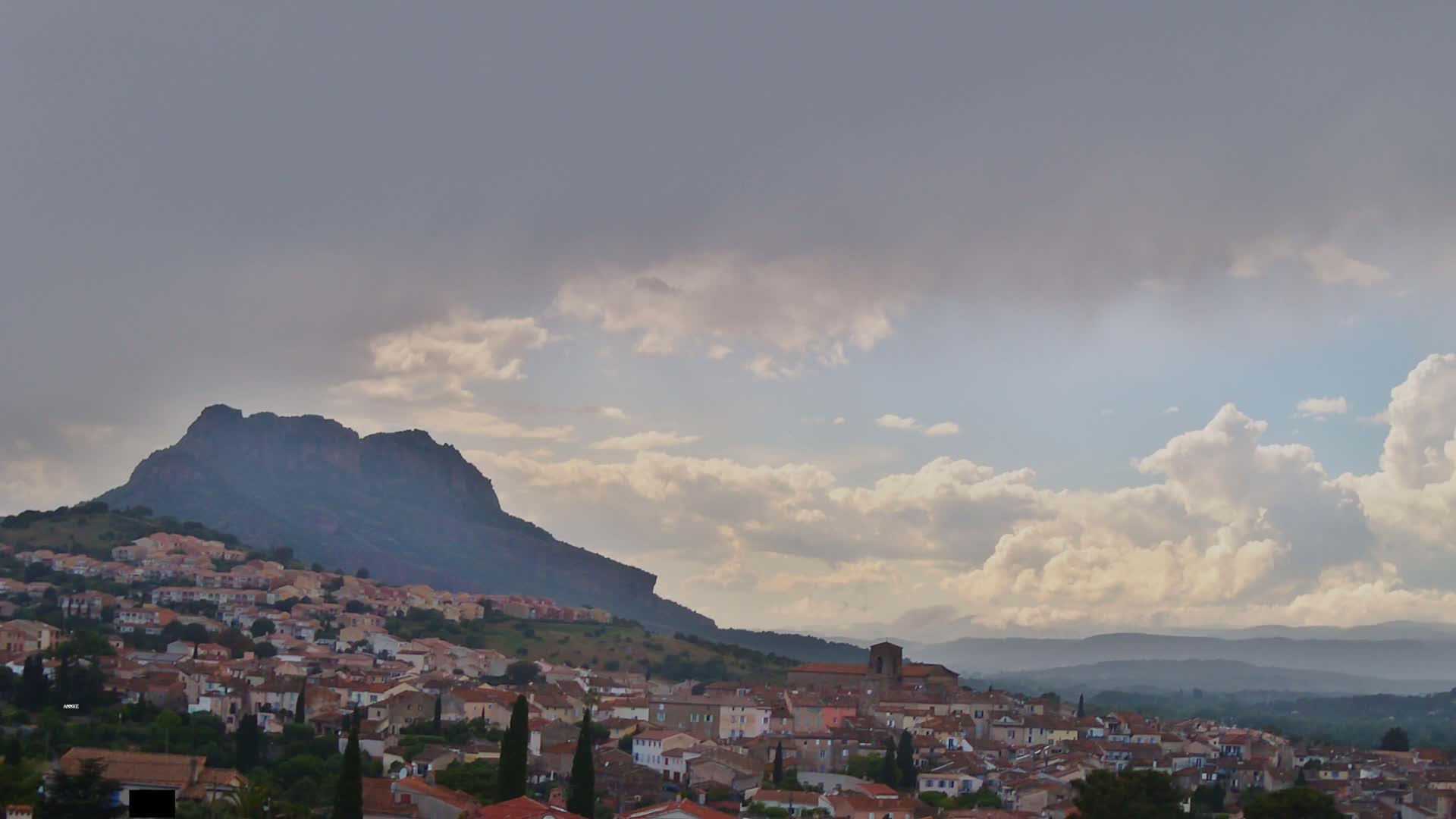 A small town with red-tiled roofs nestles against a large, dark mountain under a partly cloudy sky.