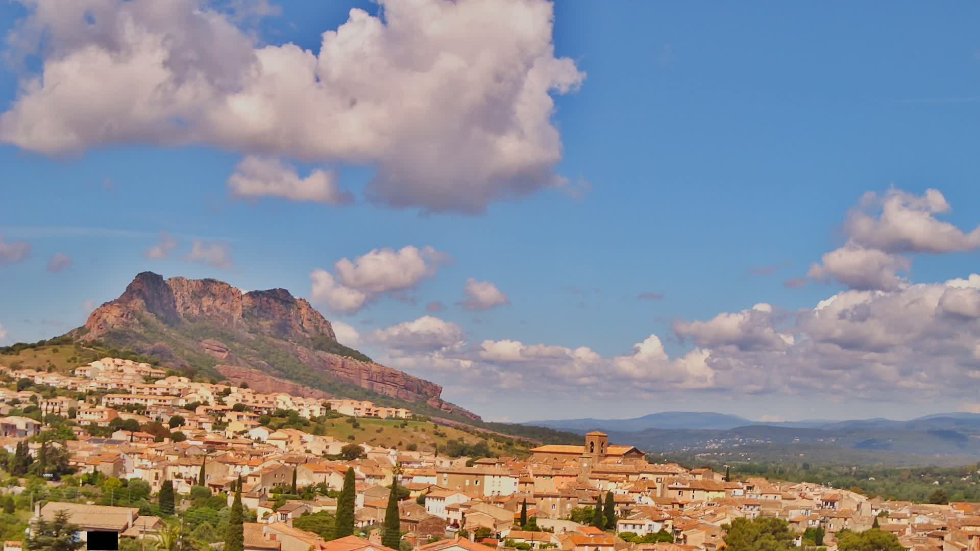 A hillside village of tan-colored buildings nestles against a large reddish-brown mountain under a partly cloudy blue sky.
