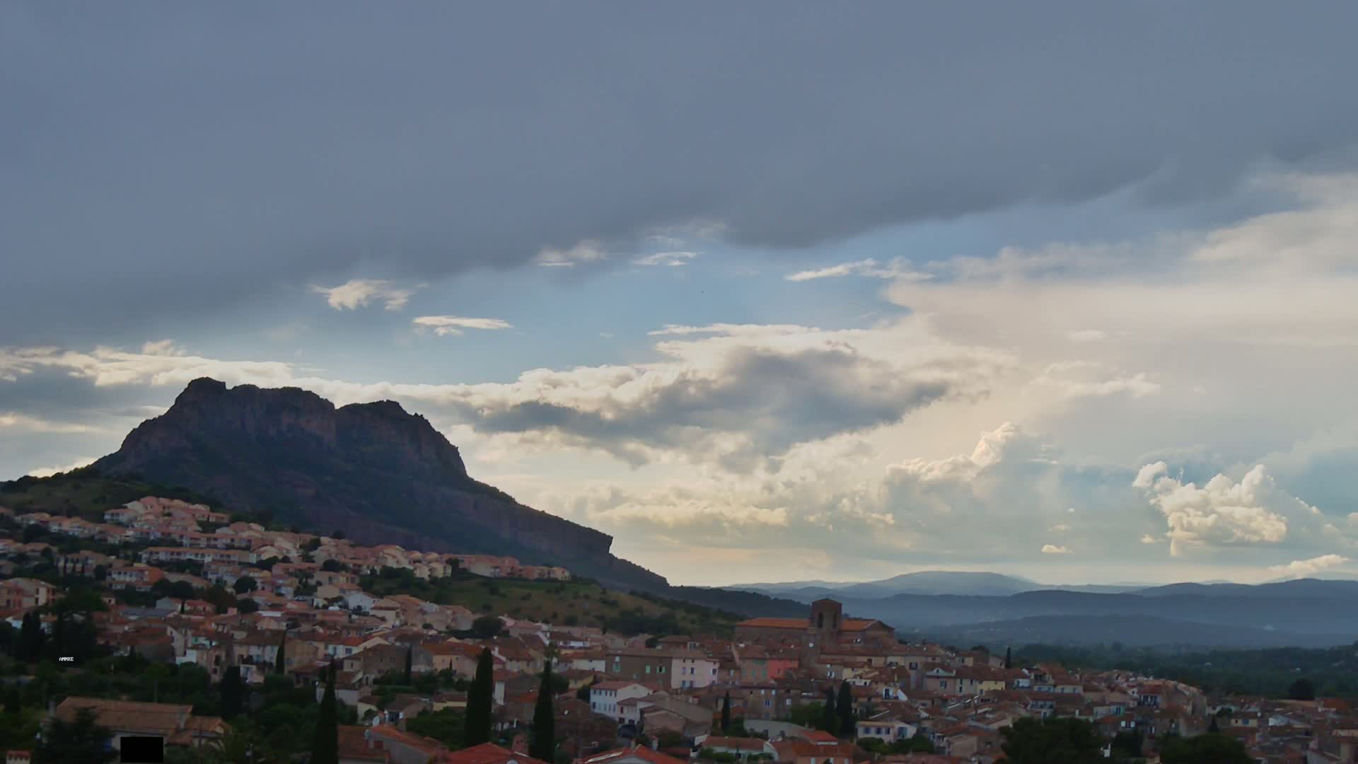 A village of red-roofed houses nestled on a hillside below a large, dark mountain under a partly cloudy sky.