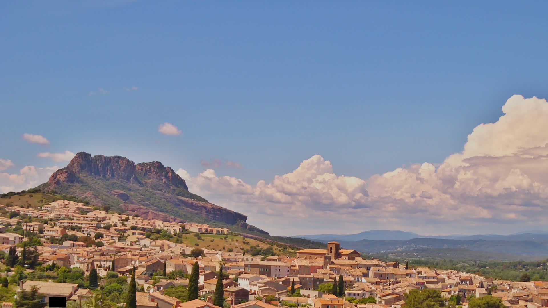 A sprawling town of tan-colored buildings nestles at the base of a large, rocky mountain under a partly cloudy blue sky.