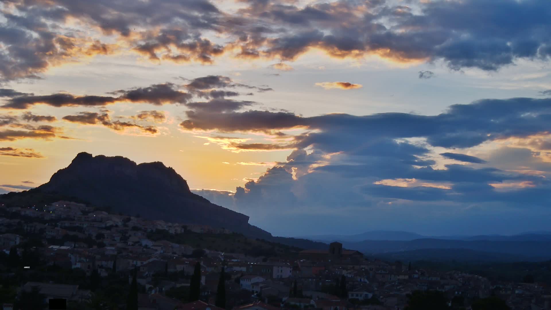 A hilltop town nestled against a silhouetted mountain at sunset under a partly cloudy sky.