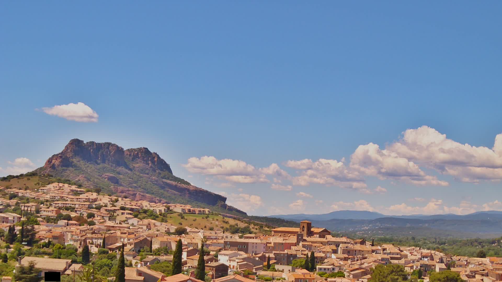 A sunny day reveals a village nestled against a rocky mountain, with fluffy white clouds scattered across a clear blue sky and distant mountains in the background.