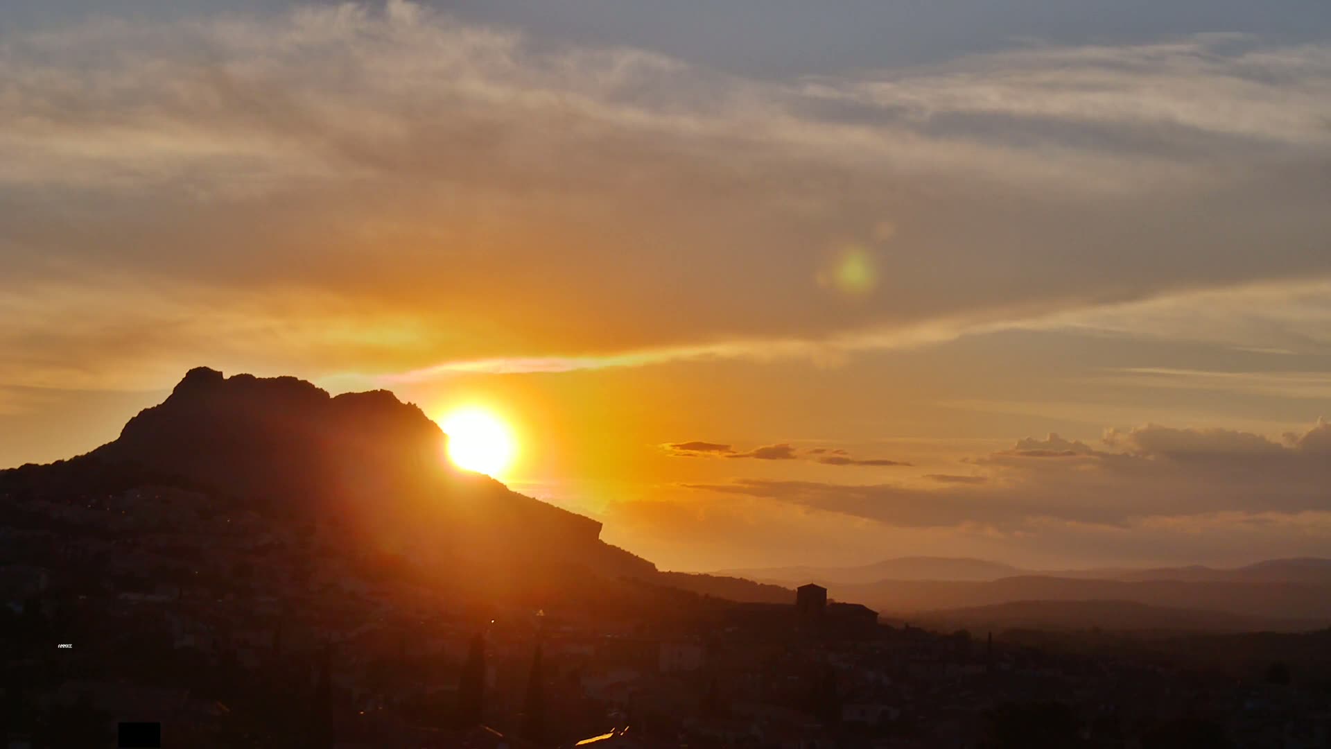 A large, dark mountain silhouetted against a vibrant sunset over a town and distant hills under a partly cloudy sky.