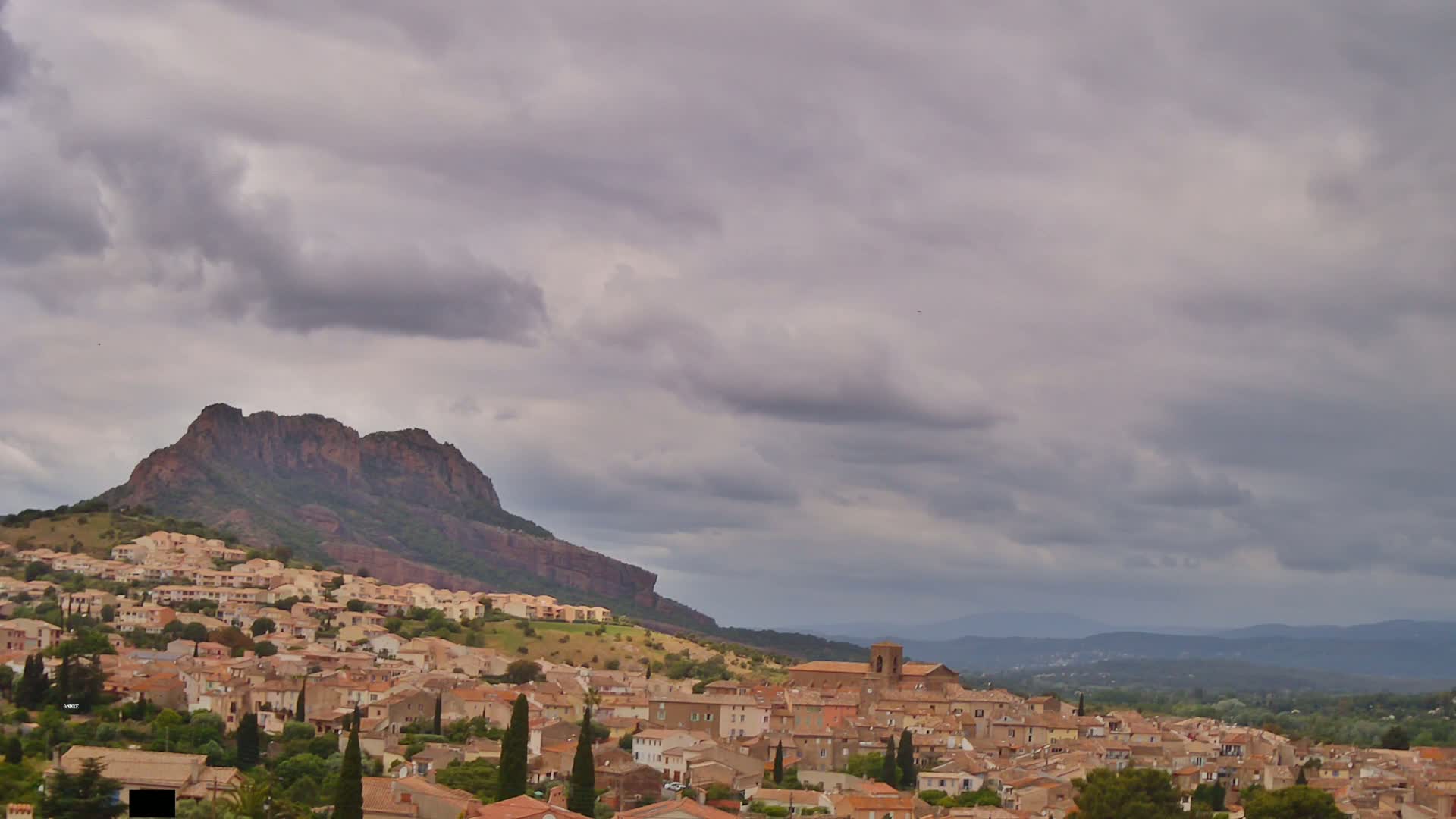 A hilltop village of tan and brown buildings nestles below a large, rocky mountain under a cloudy sky.