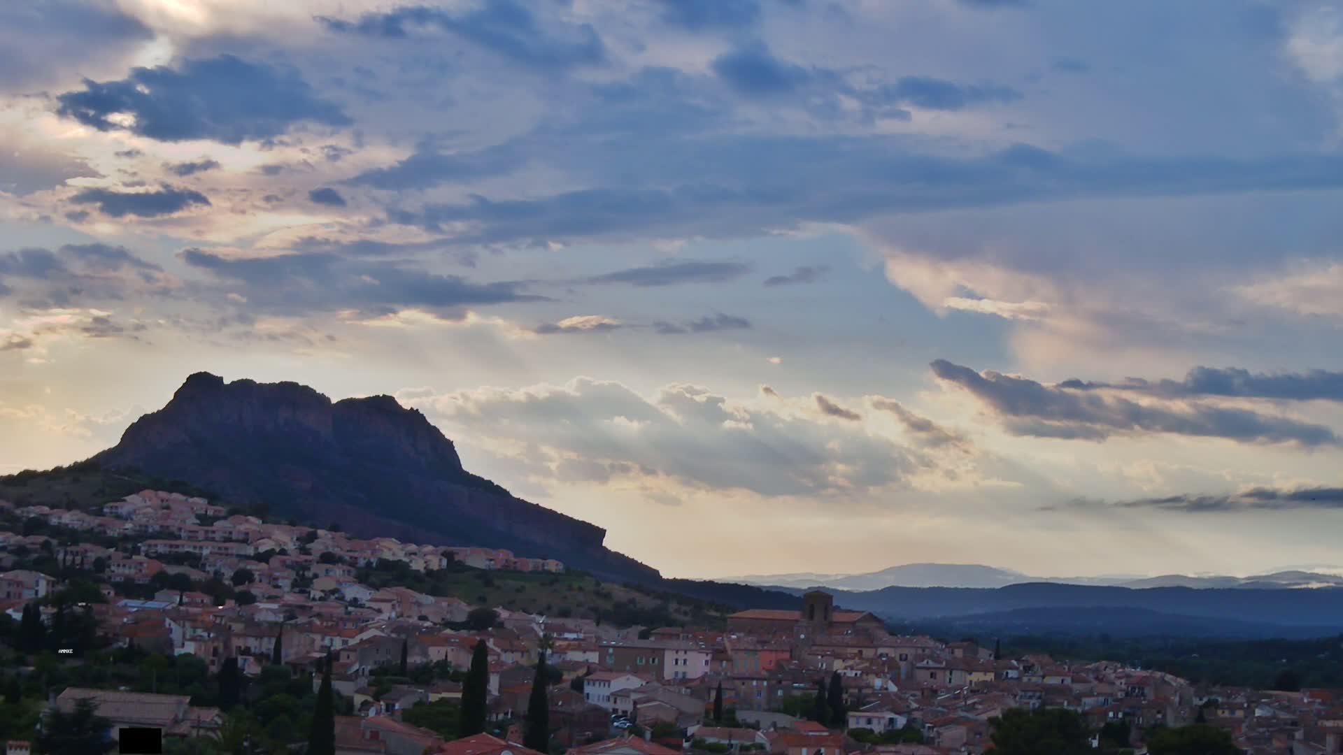 A small town nestled at the base of a large, dark mountain under a partly cloudy sky.