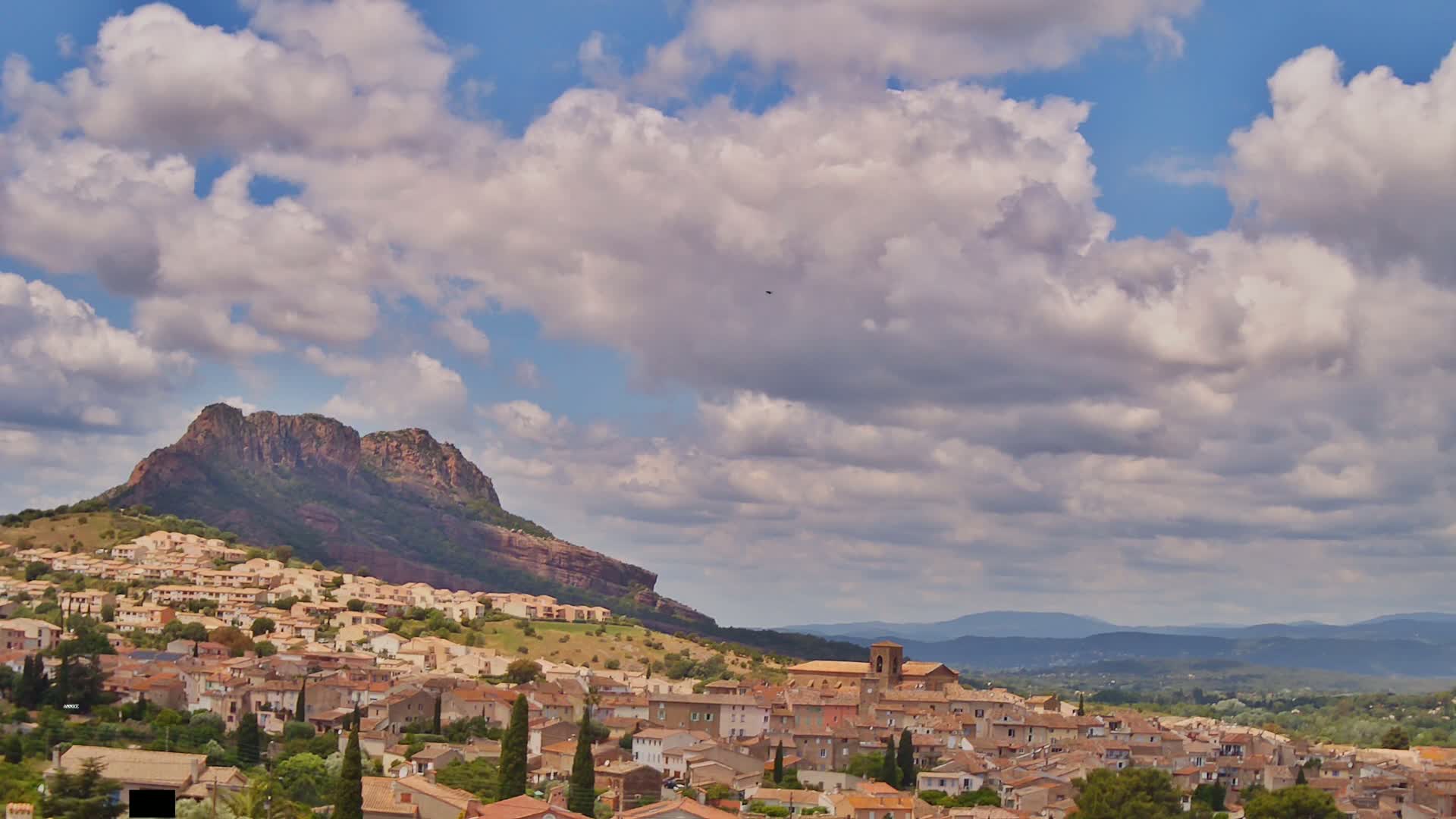 A partly cloudy sky overlooks a town nestled at the base of a large, rocky hill, with additional mountains visible in the distance.