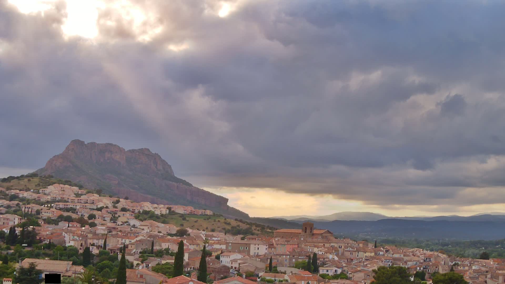 Sunbeams pierce a cloudy sky over a hillside town nestled against a large, rocky mountain.