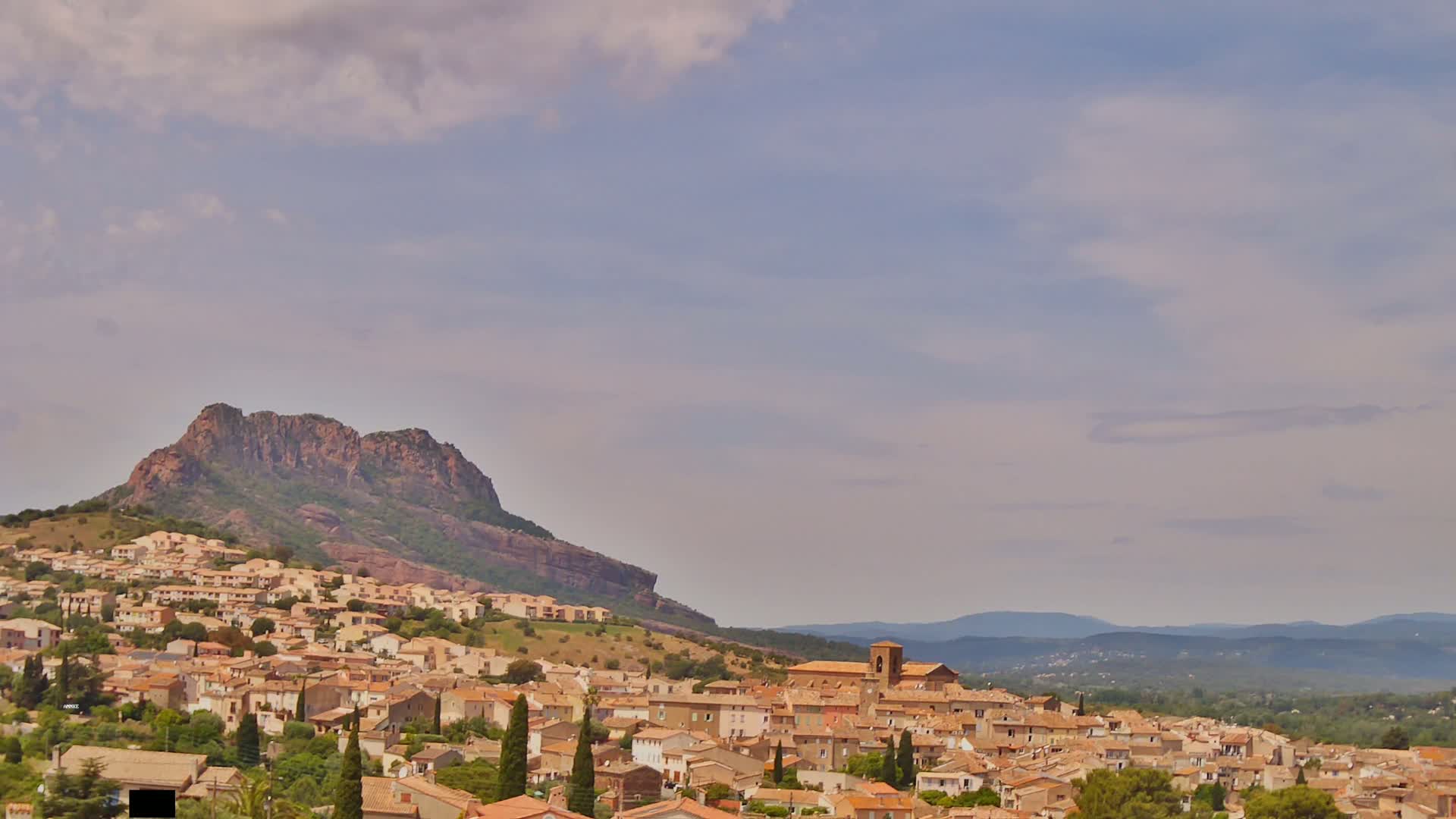 A hillside town of tan buildings nestles beneath a rocky mountain under a partly cloudy sky.