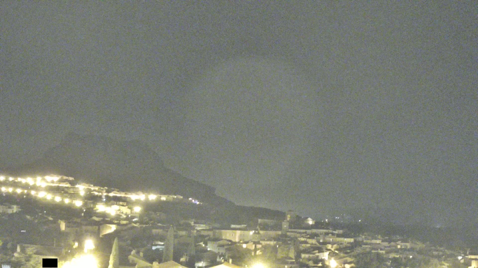 A nighttime view of a hilltop town illuminated by streetlights under a dark, overcast sky.