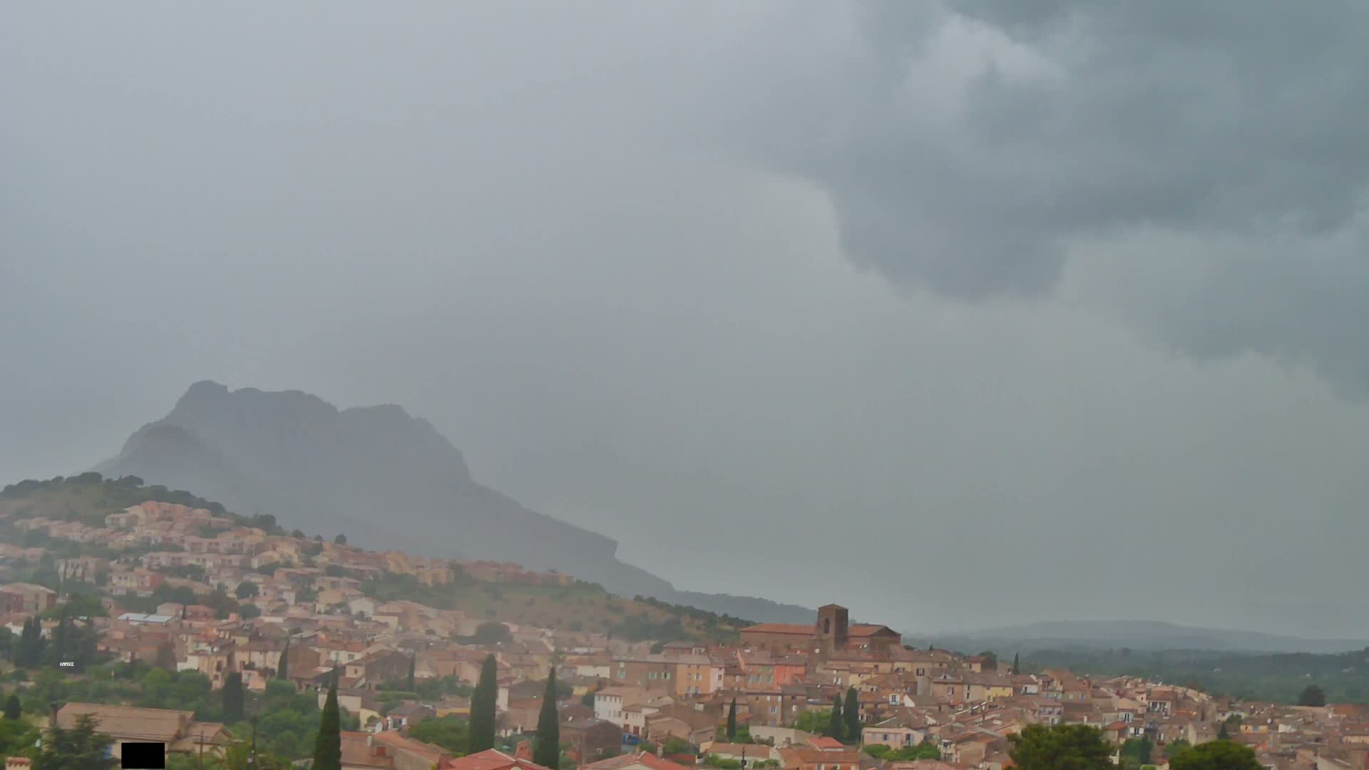 A hillside town with terracotta roofs sits under a dark, overcast sky, with a large mountain in the background.
