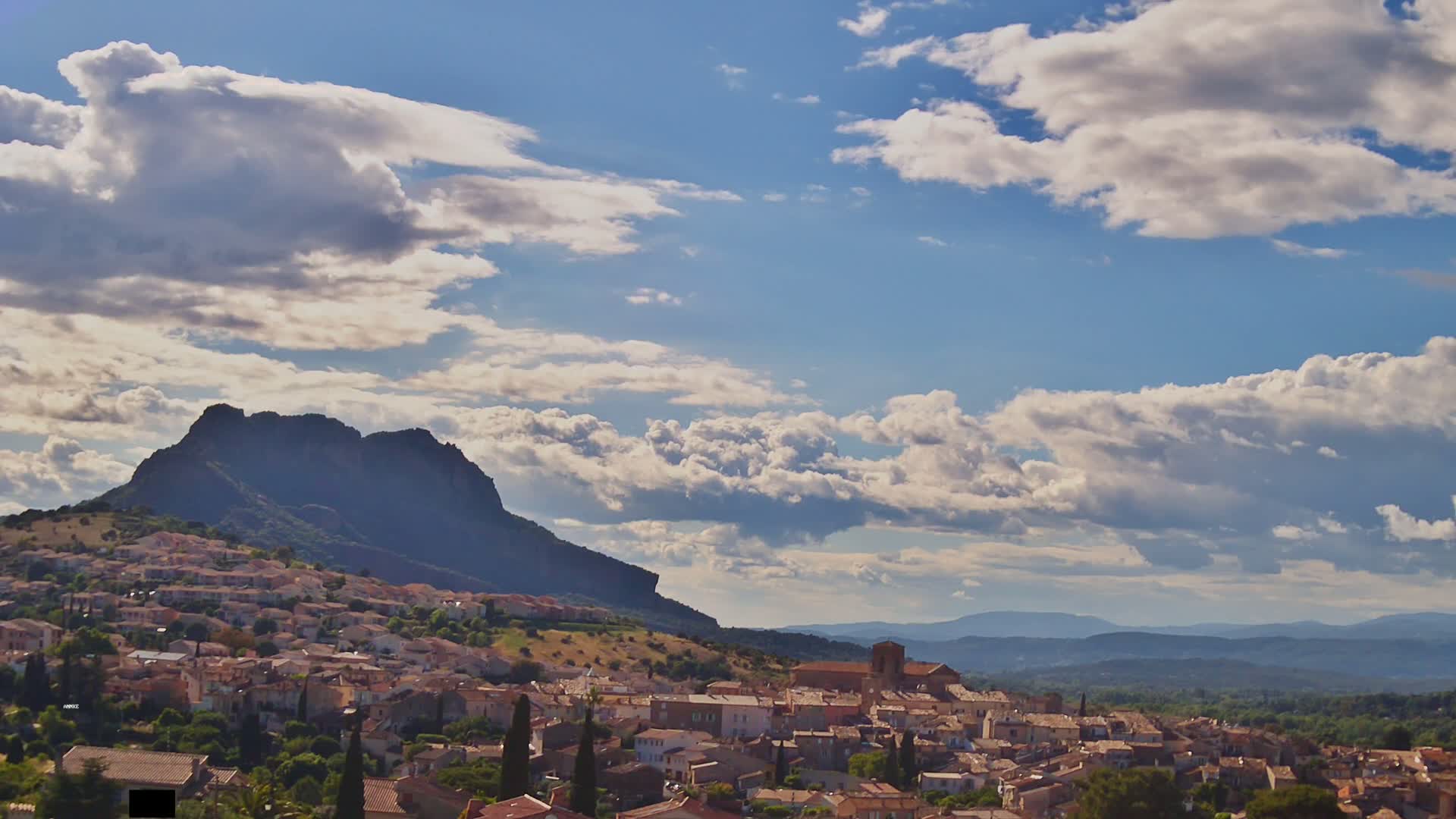 A small town nestled against a large, dark mountain under a partly cloudy sky.