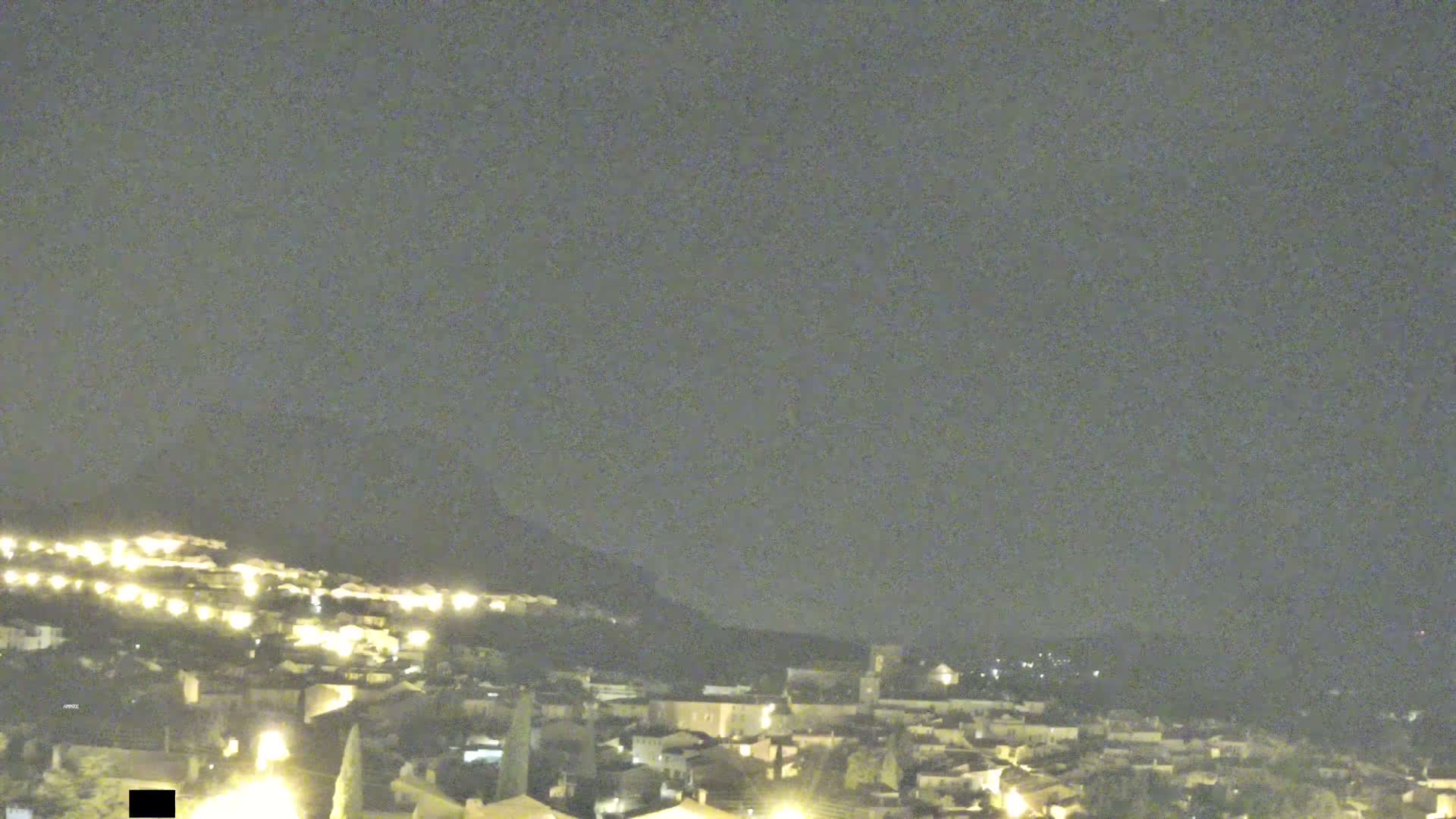 A nighttime long shot of a town nestled on a hill under a cloudy sky.
