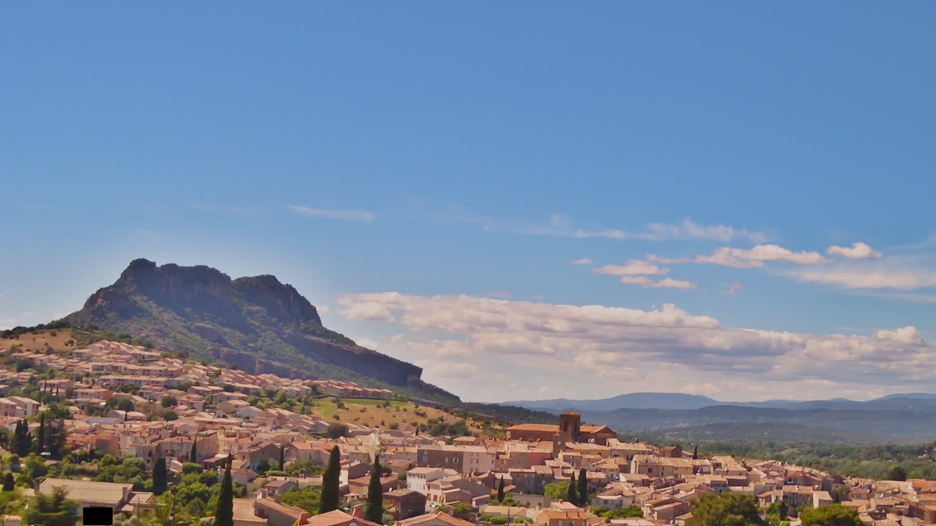 A small town with light brown buildings nestles at the base of a large, dark-colored mountain under a mostly clear blue sky with scattered puffy clouds.