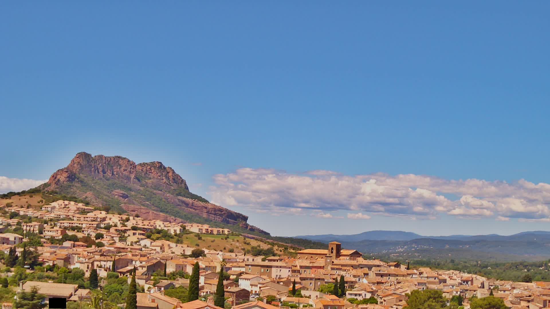A town of tan-colored buildings nestled against a rocky, reddish-brown hill under a mostly clear blue sky with a few fluffy white clouds.