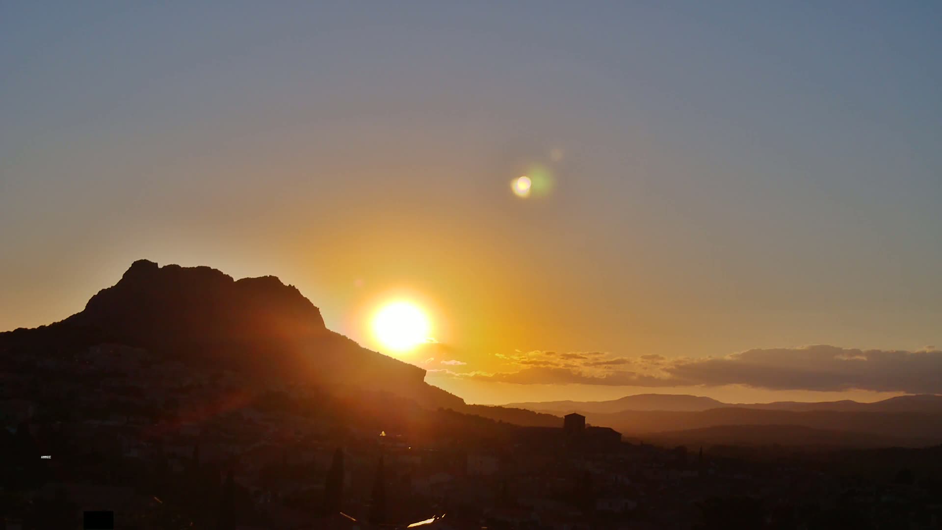A large, dark mountain silhouetted against a vibrant sunset over a town, with a partly cloudy sky.