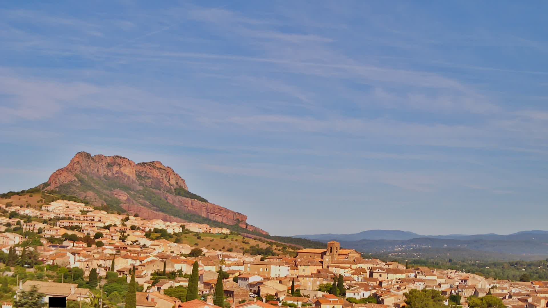 A hilltop town with tan-colored buildings and reddish-brown mountains stretches beneath a clear, light blue sky.