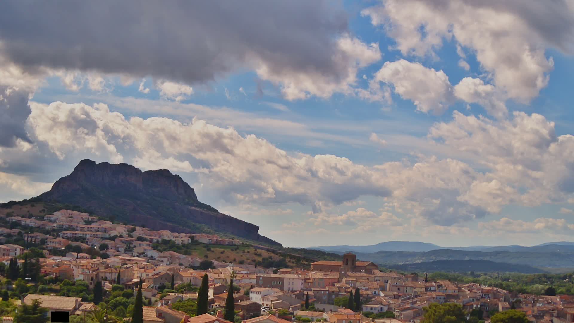 A partly cloudy sky overlooks a village nestled at the base of a large, dark-colored mountain, with rolling hills visible in the distance.
