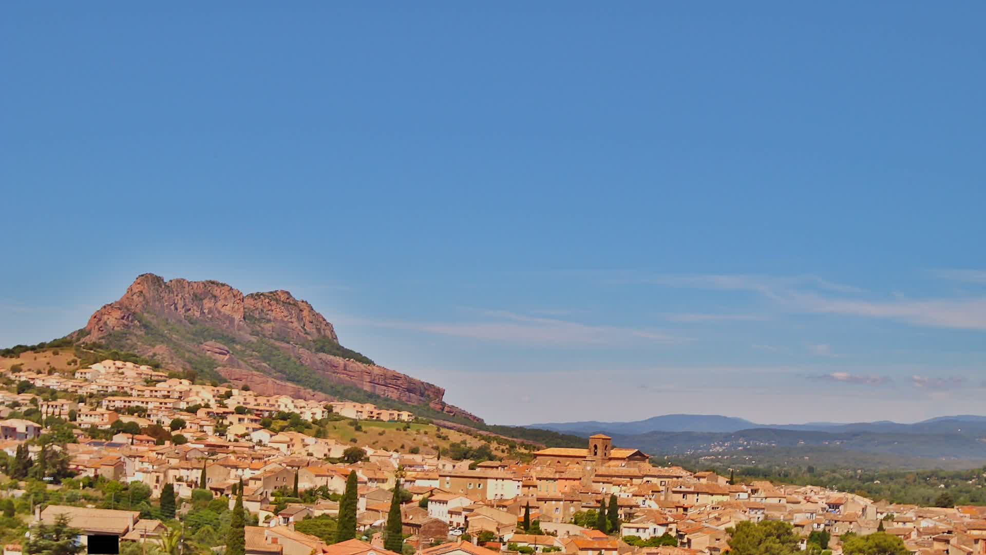 A small town of tan buildings nestles at the base of a large, reddish-brown mountain under a clear blue sky.