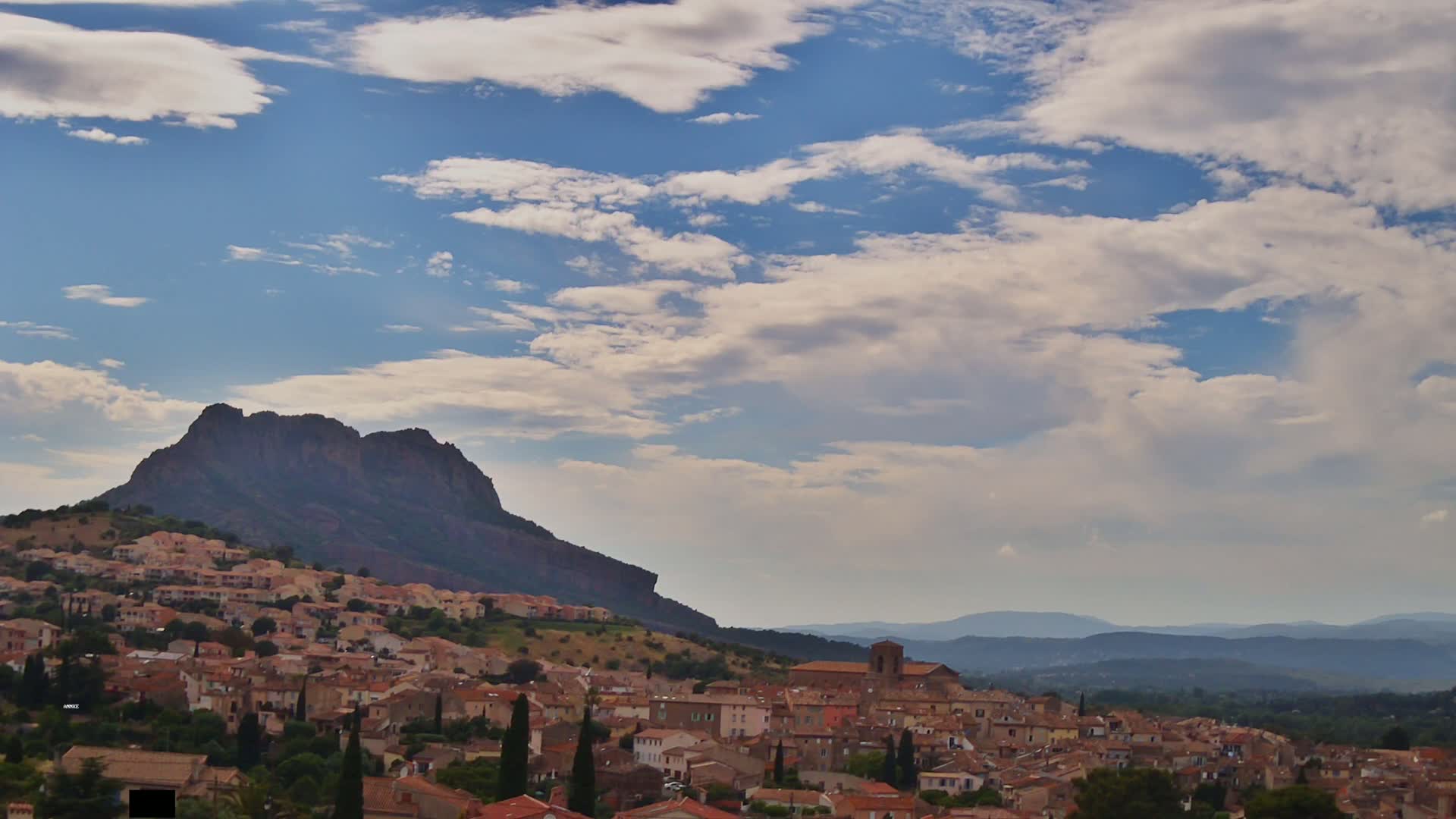 A partly cloudy sky overlooks a hillside town nestled against a large, dark mountain range, extending to distant, hazy mountains.
