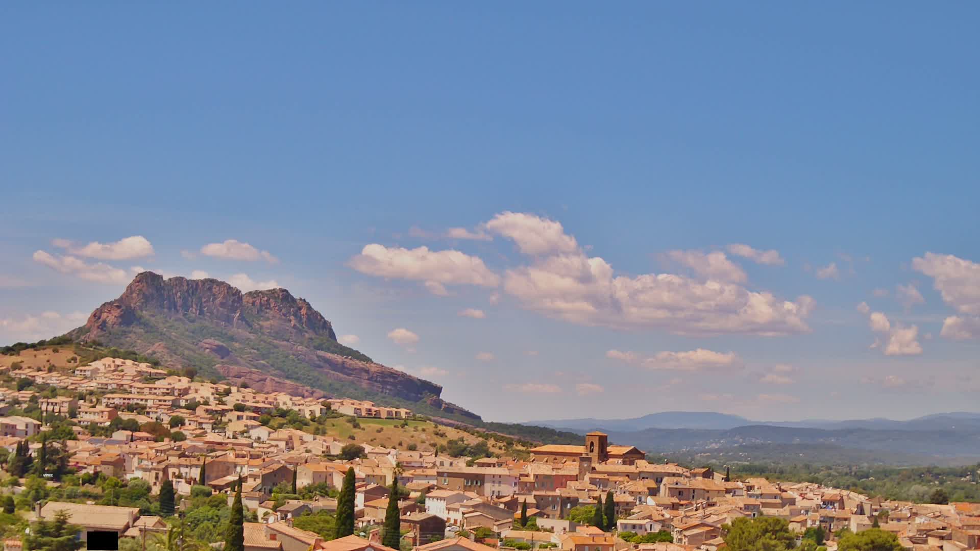A hilltop village with tan buildings is nestled beneath a rocky mountain under a mostly sunny sky with scattered puffy clouds.