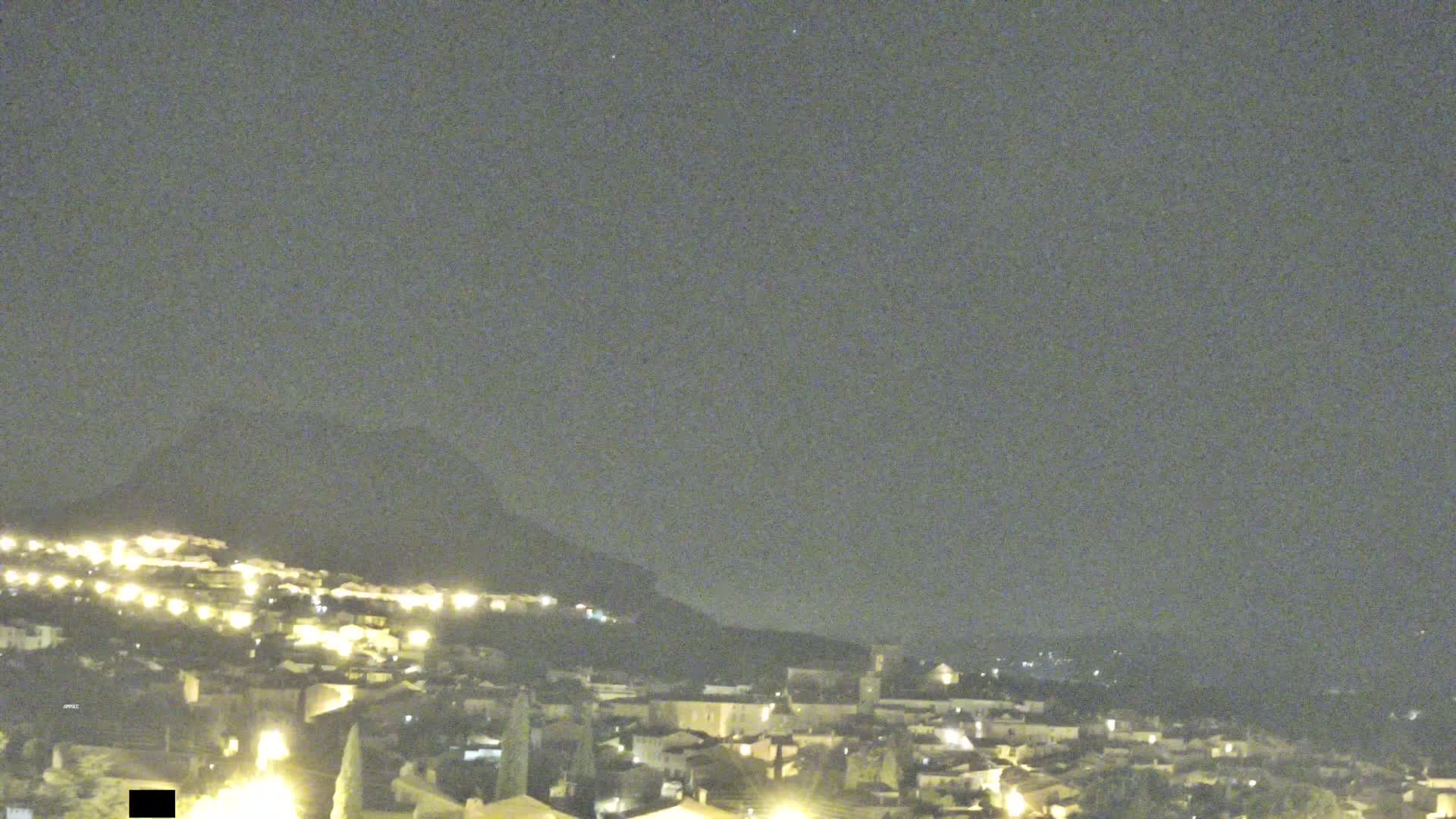 A nighttime view shows a hilltop town illuminated by many lights under a dark, clear sky.