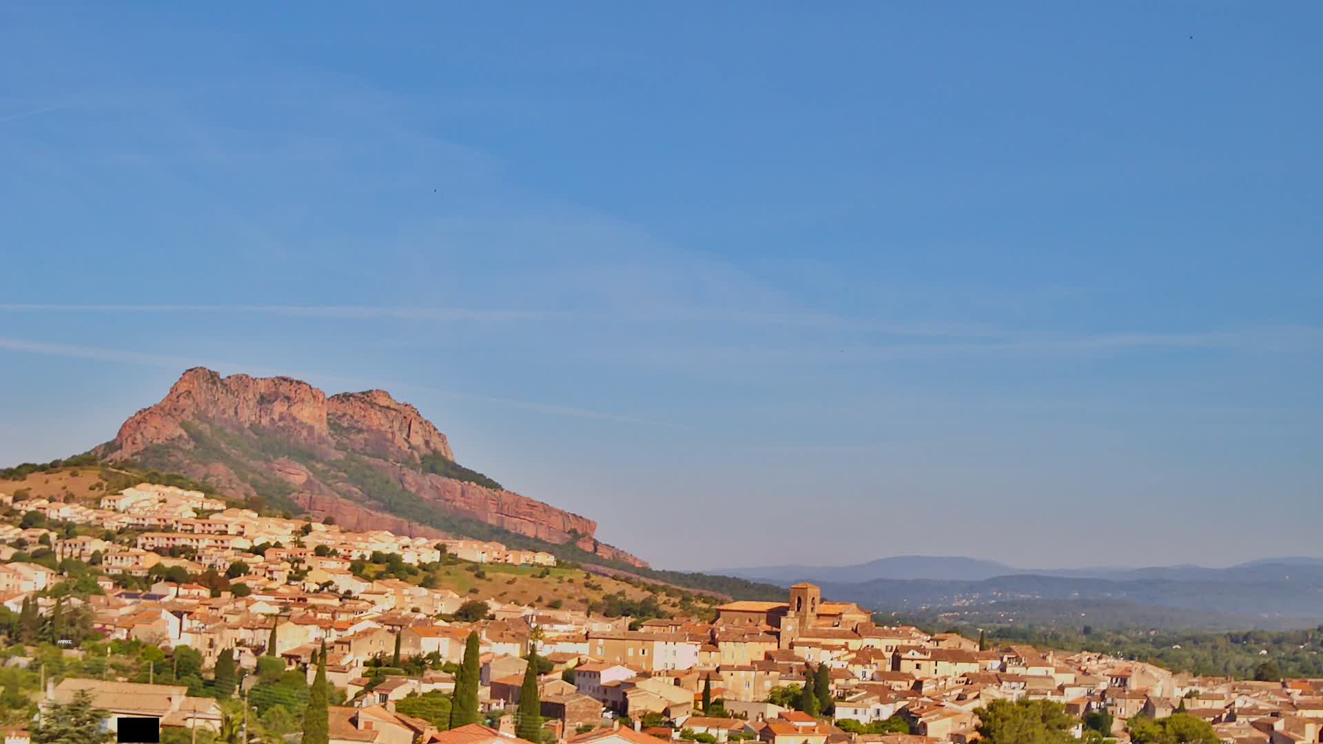 A town of light-brown buildings nestled against a large, reddish-brown mountain under a clear blue sky.