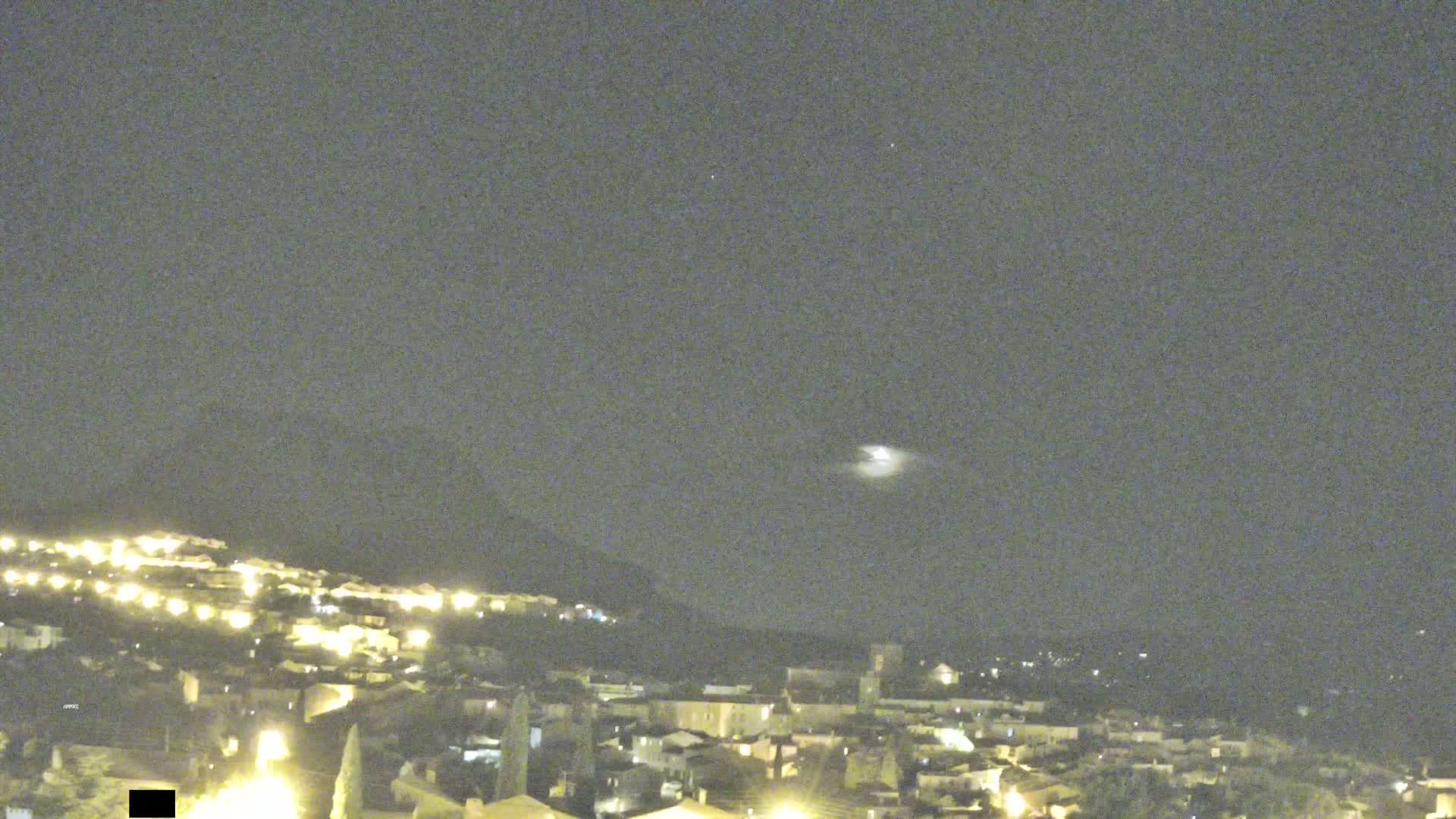 A nighttime view of a town nestled on a hillside under a mostly clear sky, with a bright moon or planet visible in the distance.