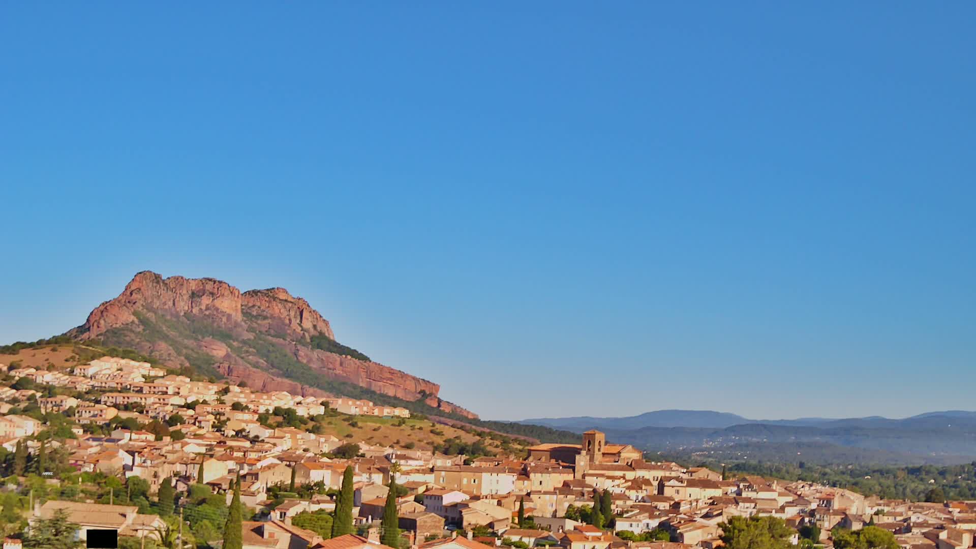 A sprawling village of tan and brown buildings nestles at the base of a large, reddish-brown mountain under a clear blue sky.
