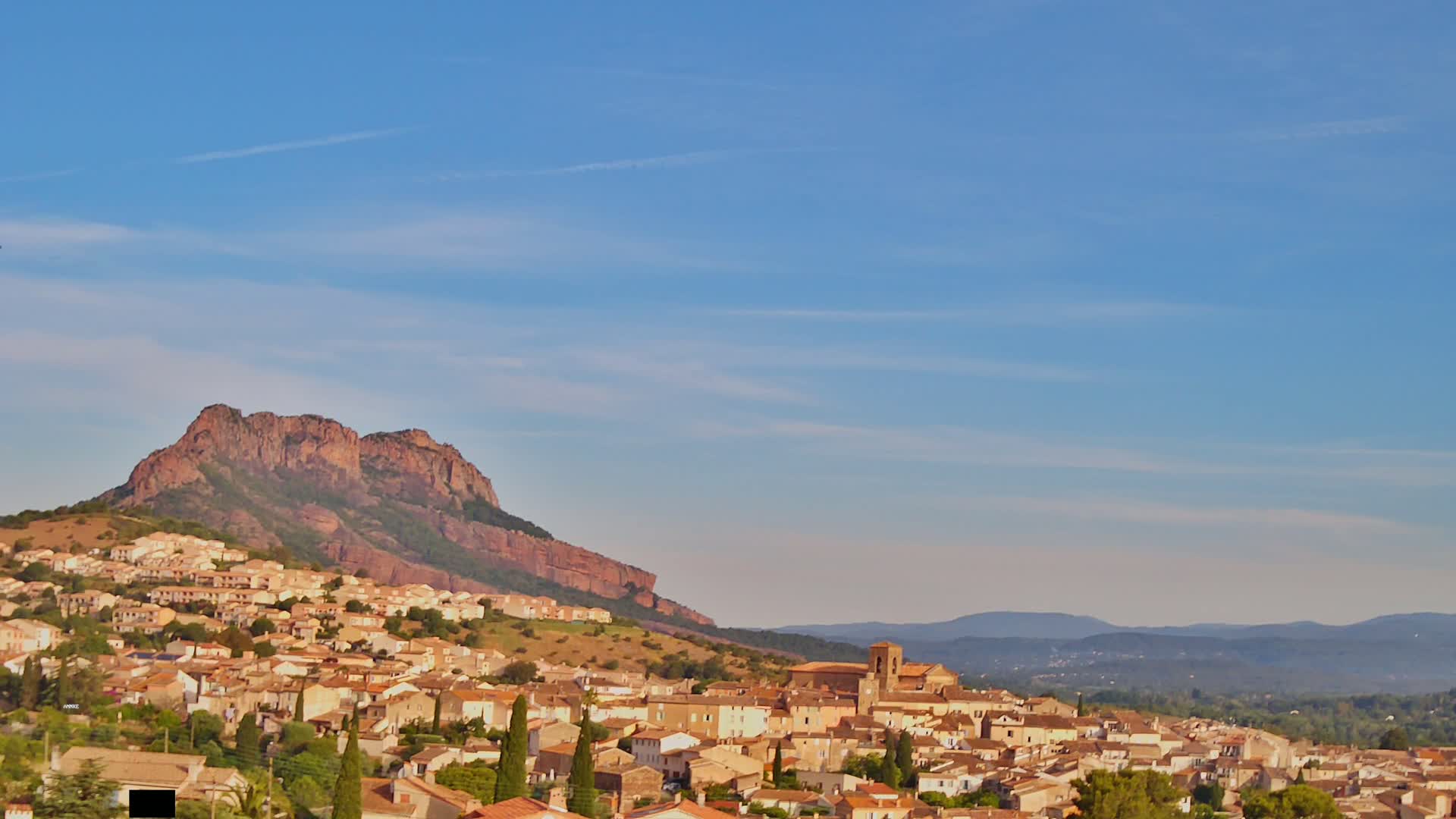 A small town of tan buildings nestled against a large, reddish-brown mountain under a clear, light-blue sky.