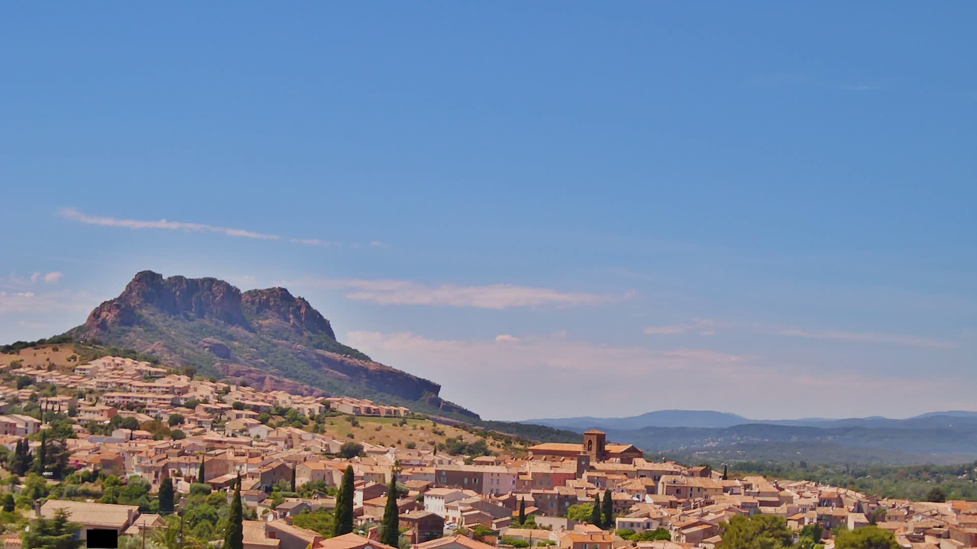 A sprawling town of tan-colored buildings nestles against a rocky, tree-covered hill under a mostly clear blue sky.