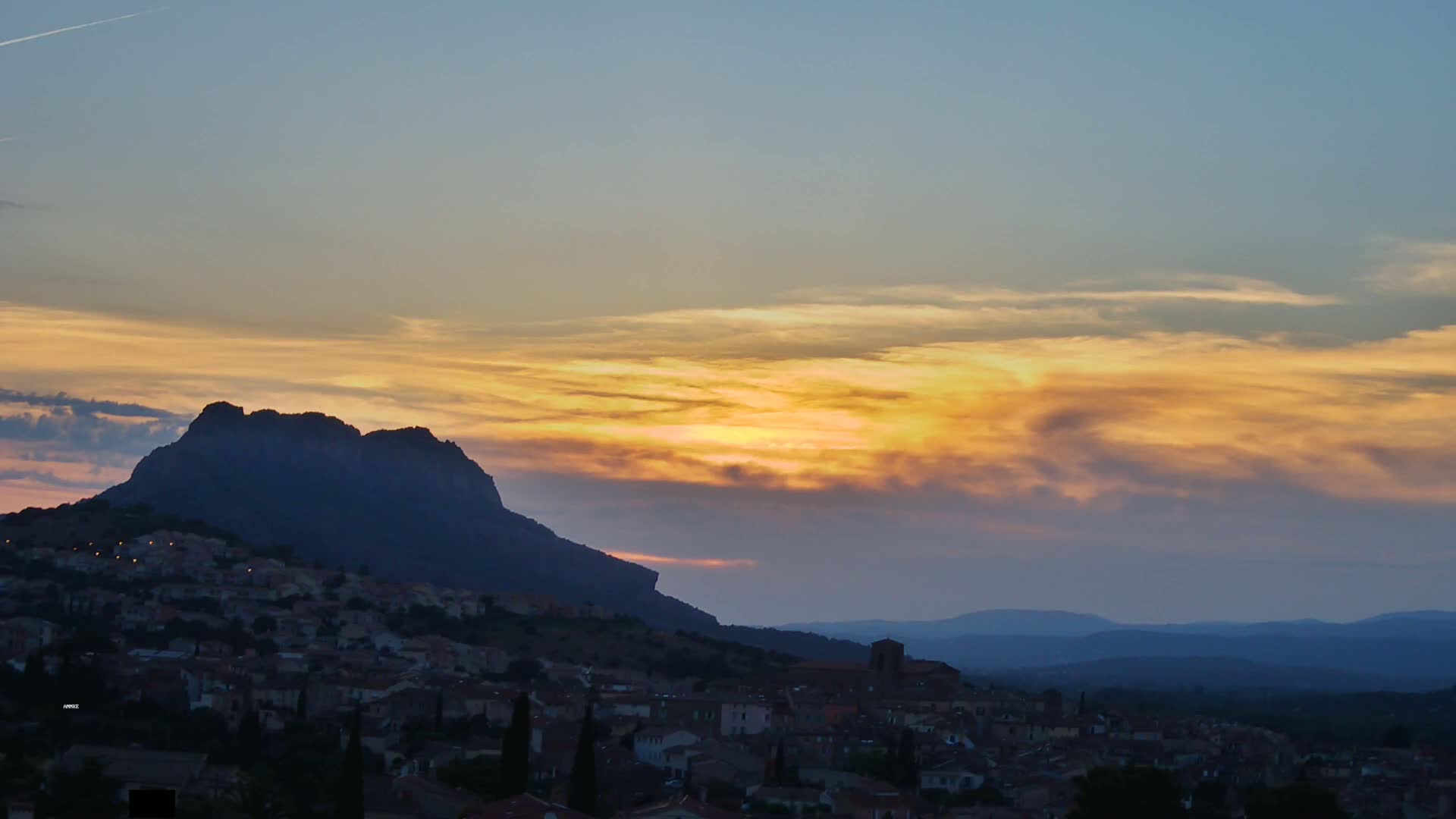 A small town nestled at the foot of a large, dark mountain is silhouetted against a sunset sky filled with golden and purple clouds.