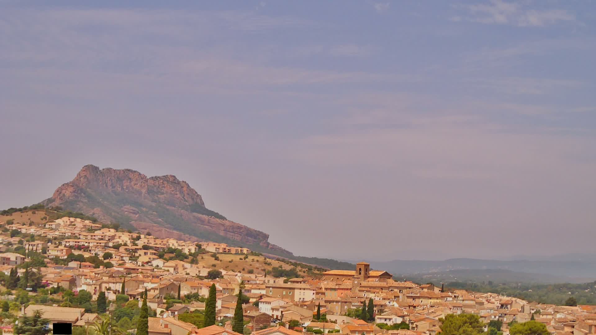 A hillside town with tan-colored buildings nestles below a large, rocky mountain under a hazy, light-blue sky.