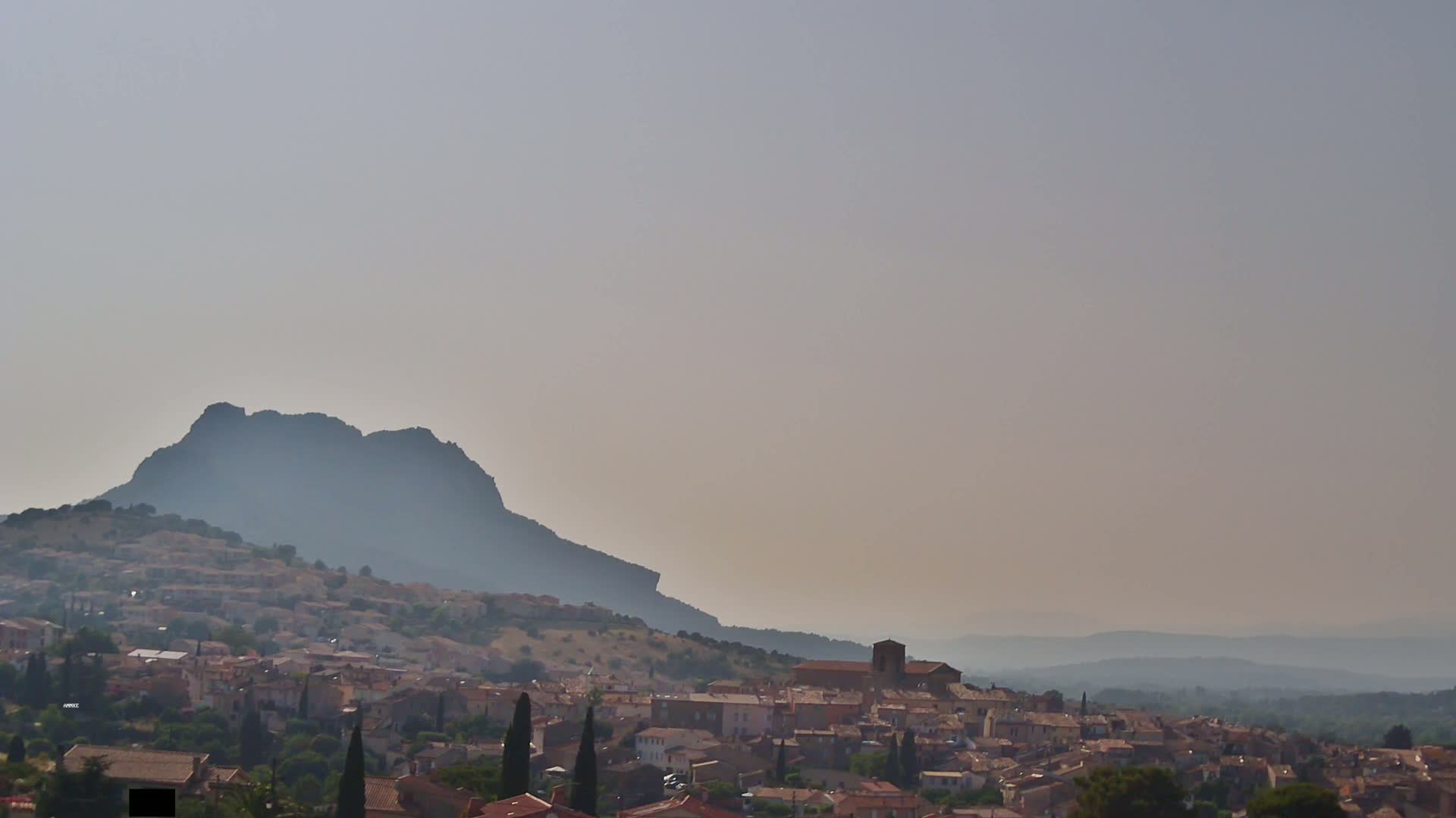 A hazy, sunlit view shows a village nestled at the base of a large, dark mountain, with rolling hills extending into the distance.