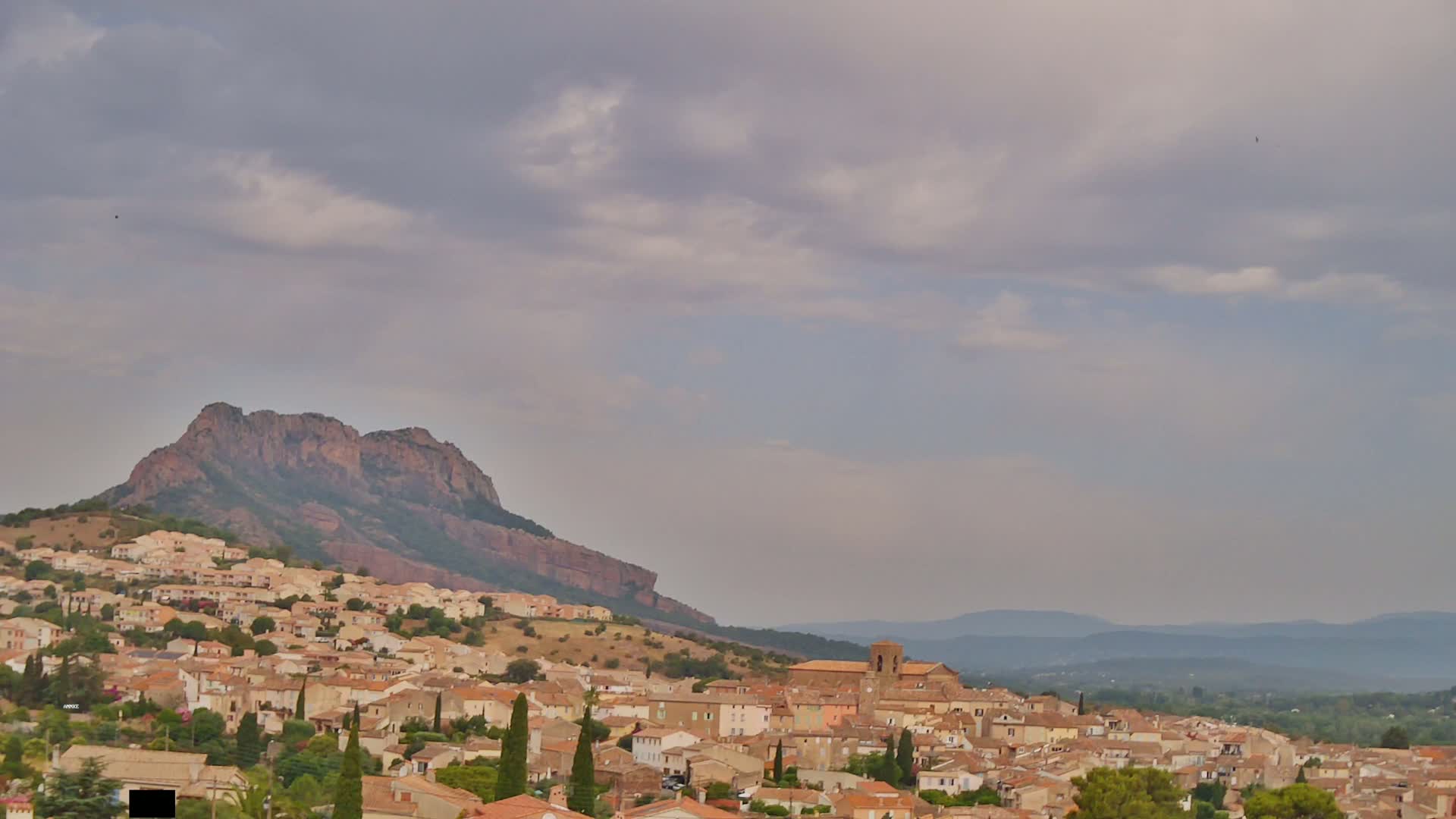 A town of tan, low buildings clusters on a hillside beneath a large, reddish-brown mountain under a mostly cloudy sky.