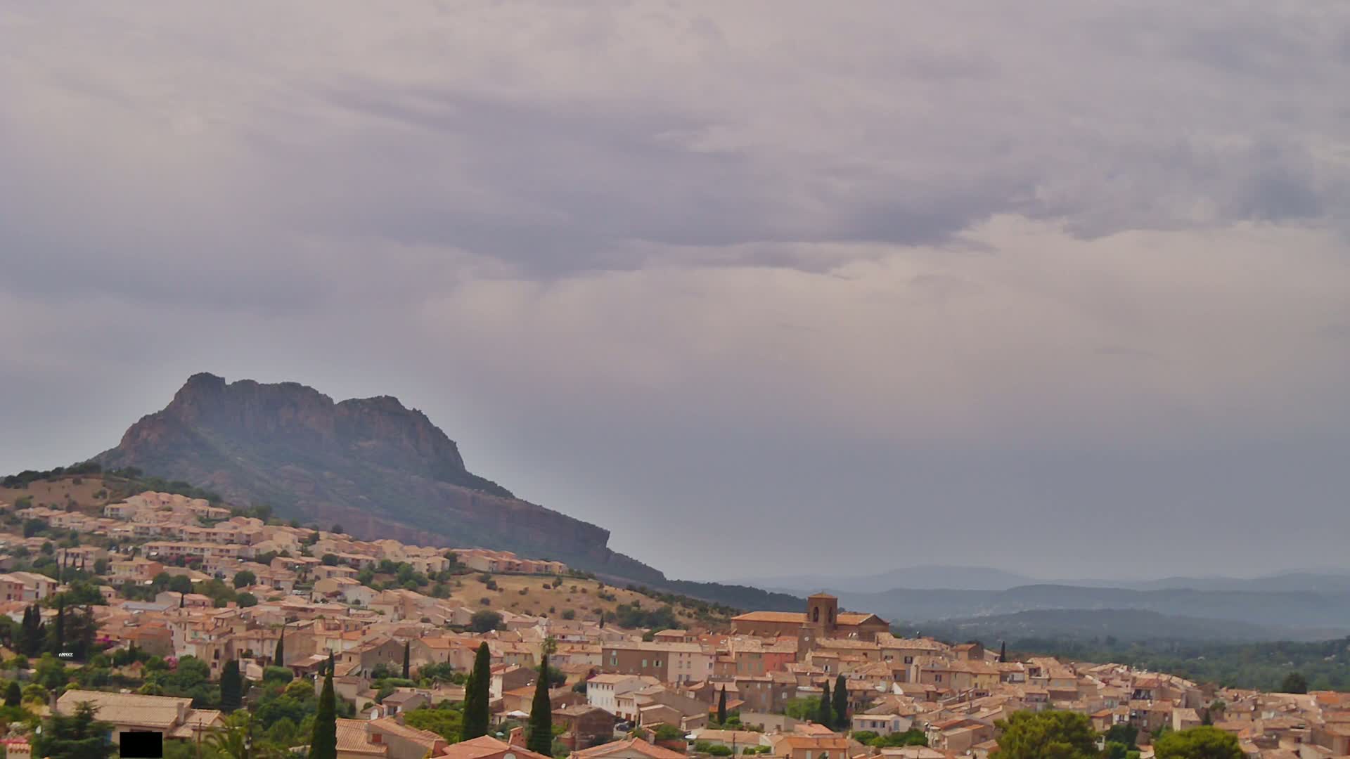 A small town of tan-colored buildings nestled on a hillside beneath a large, dark mountain under an overcast sky.