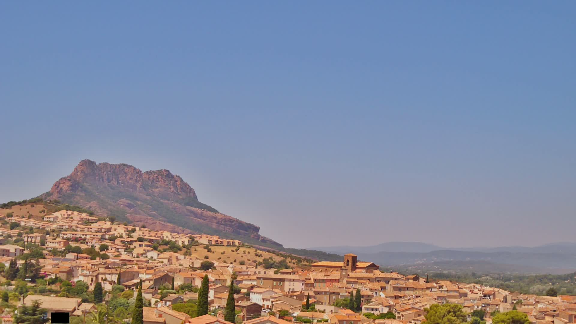 A hilltop town of tan and brown buildings stretches across a hillside beneath a clear blue sky, with a large, reddish-brown rocky mountain in the background.