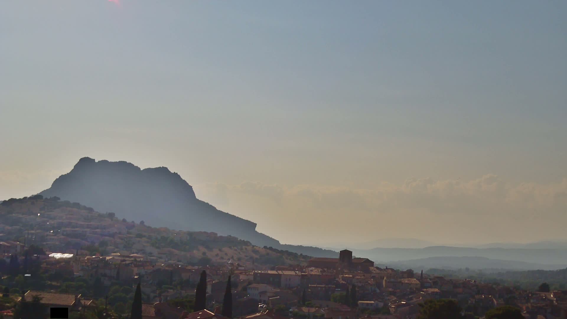 A hazy, sunlit landscape shows a hilltop town nestled against a large, dark mountain, with rolling hills extending to the horizon.