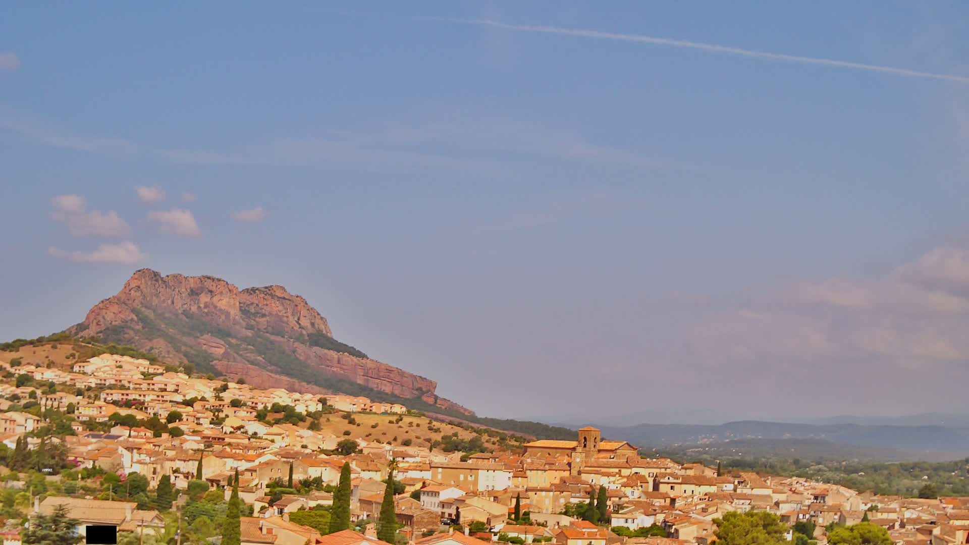 A tan-colored village with red-tiled roofs nestles against a large, reddish-brown hill under a mostly clear, light-blue sky.