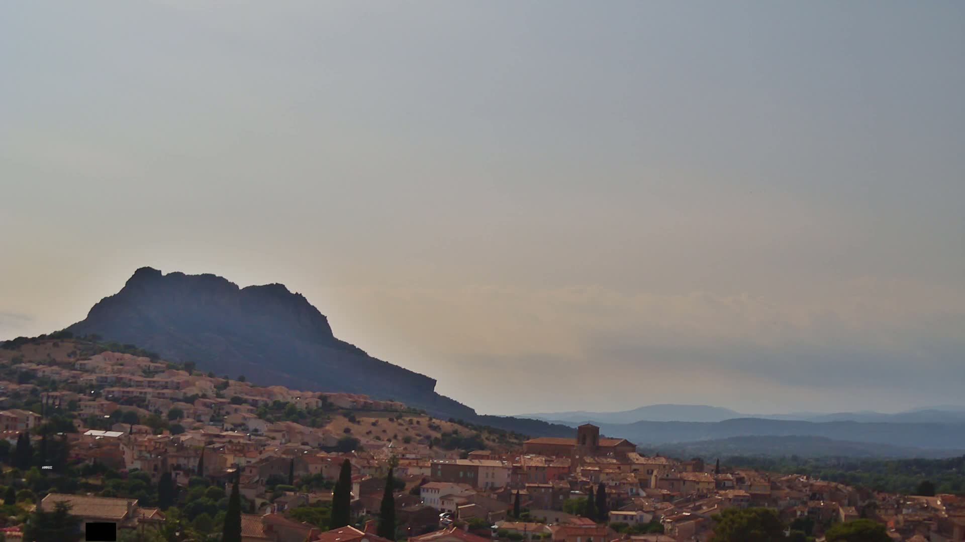 A hillside town with tan buildings nestles beneath a large, dark mountain under a hazy sky.
