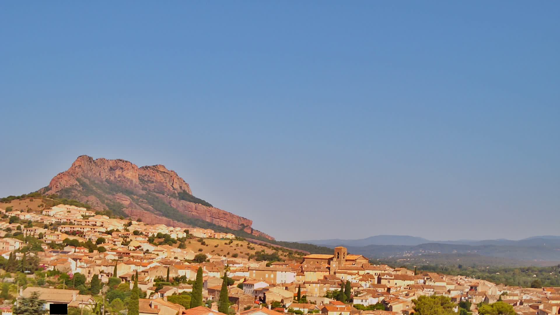 A sprawling town of tan and terracotta buildings nestles against a large, reddish-brown mountain under a clear, bright blue sky.