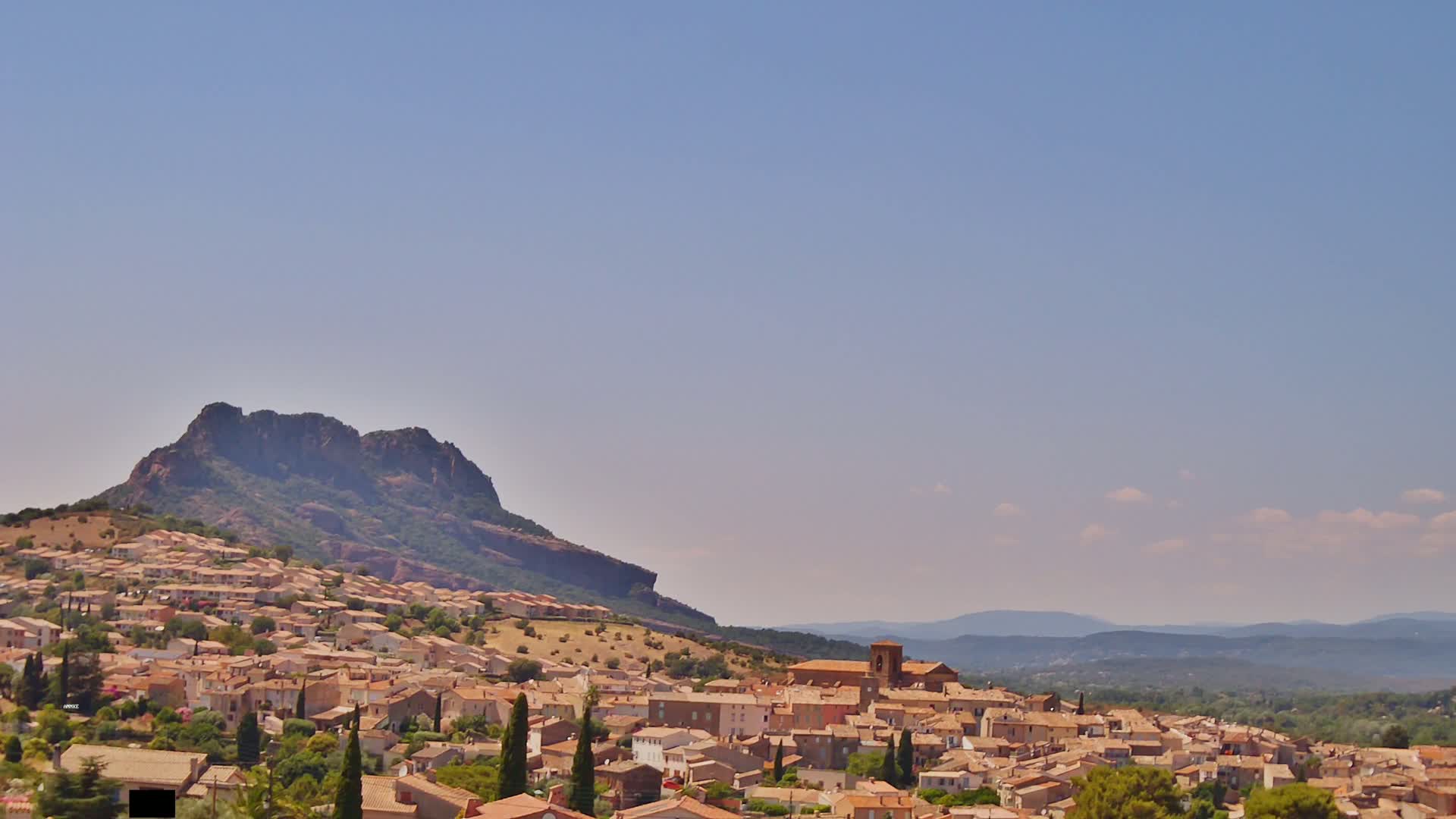 A hilltop town of tan buildings nestles against a rocky mountain under a clear, light-blue sky.