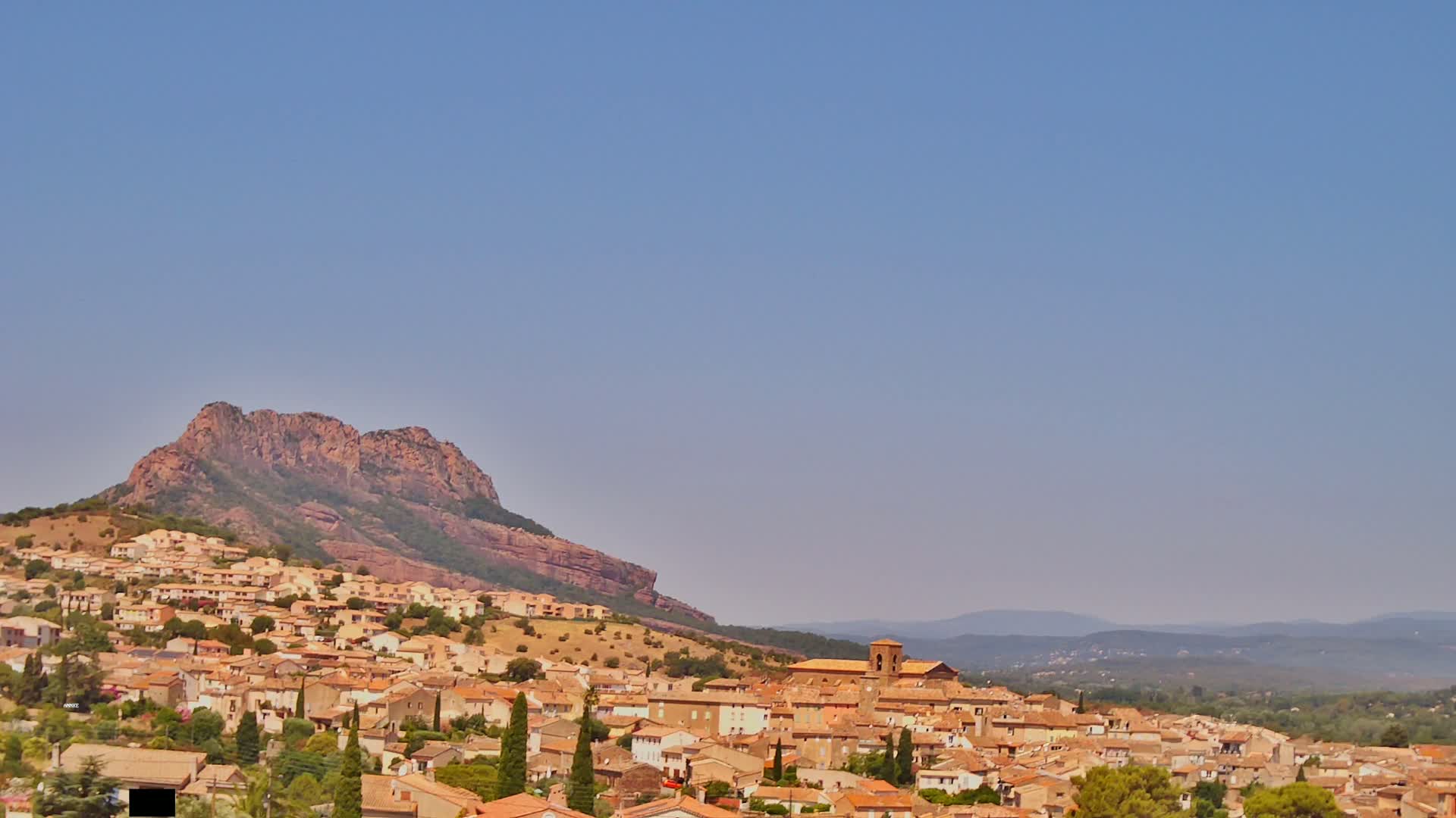A tan-colored village with red-tiled roofs sprawls across a hillside beneath a large, reddish-brown mountain under a clear, light-blue sky.