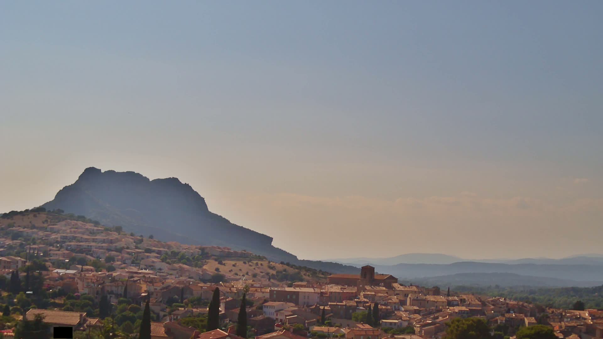 A sprawling village of tan buildings nestles against a large, dark mountain under a hazy, light-blue sky, with distant mountains visible on the horizon.