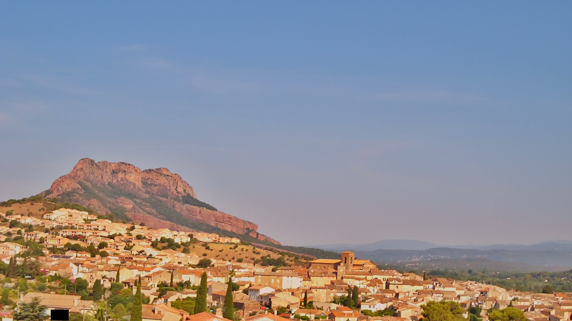 A large, tan-colored town with red-tiled roofs nestles at the base of a rocky, reddish-brown hill under a clear, pale blue sky.