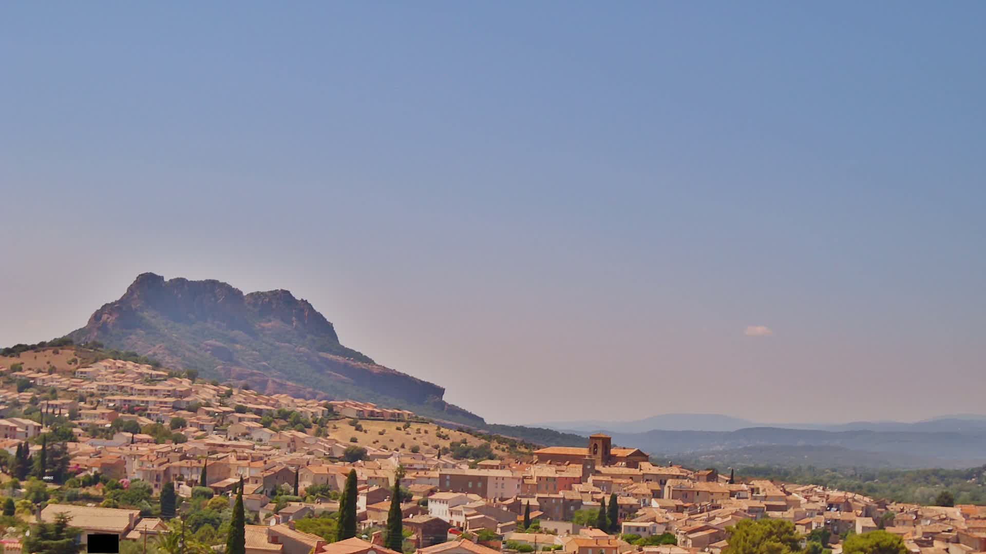 A hilltop town with tan-colored buildings sits below a large, dark mountain under a clear, light-blue sky.