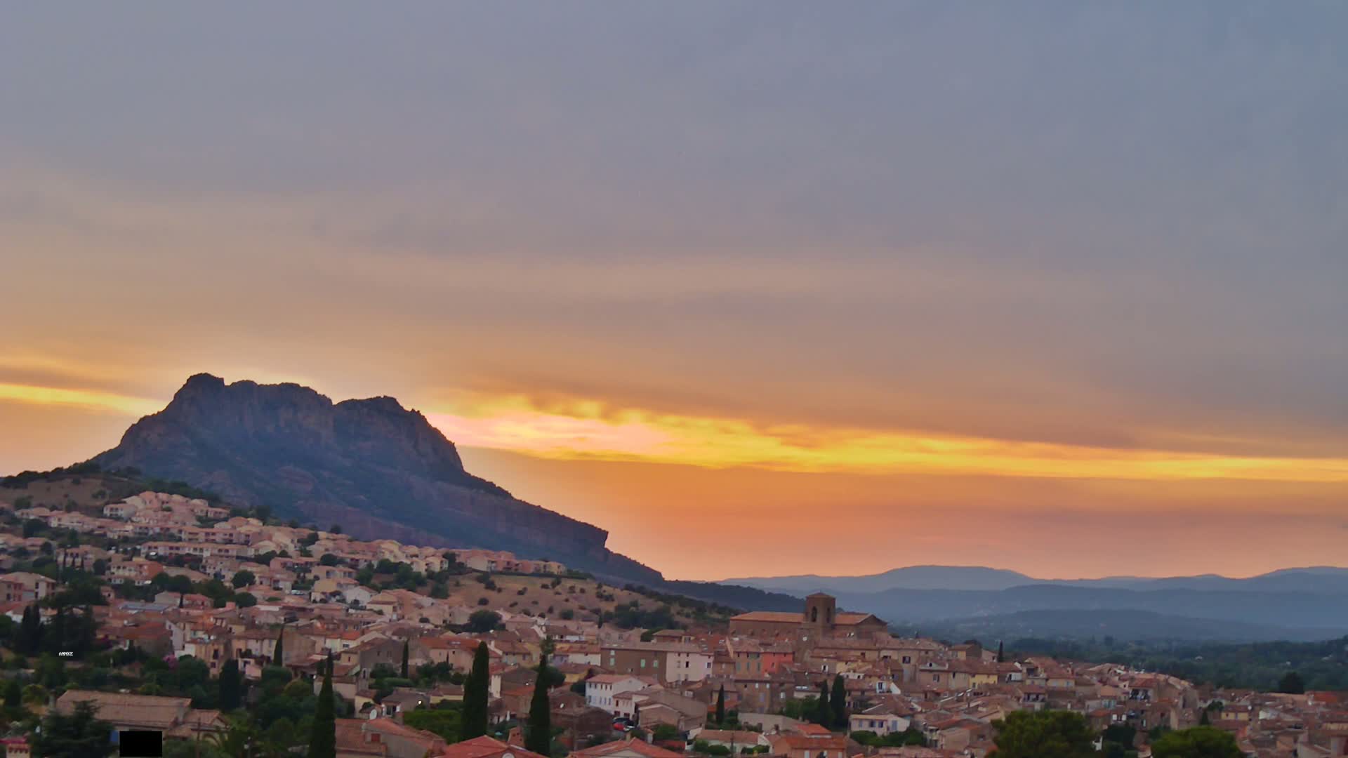 A small town nestled against a large, dark mountain at sunset, under a pale orange and yellow sky.