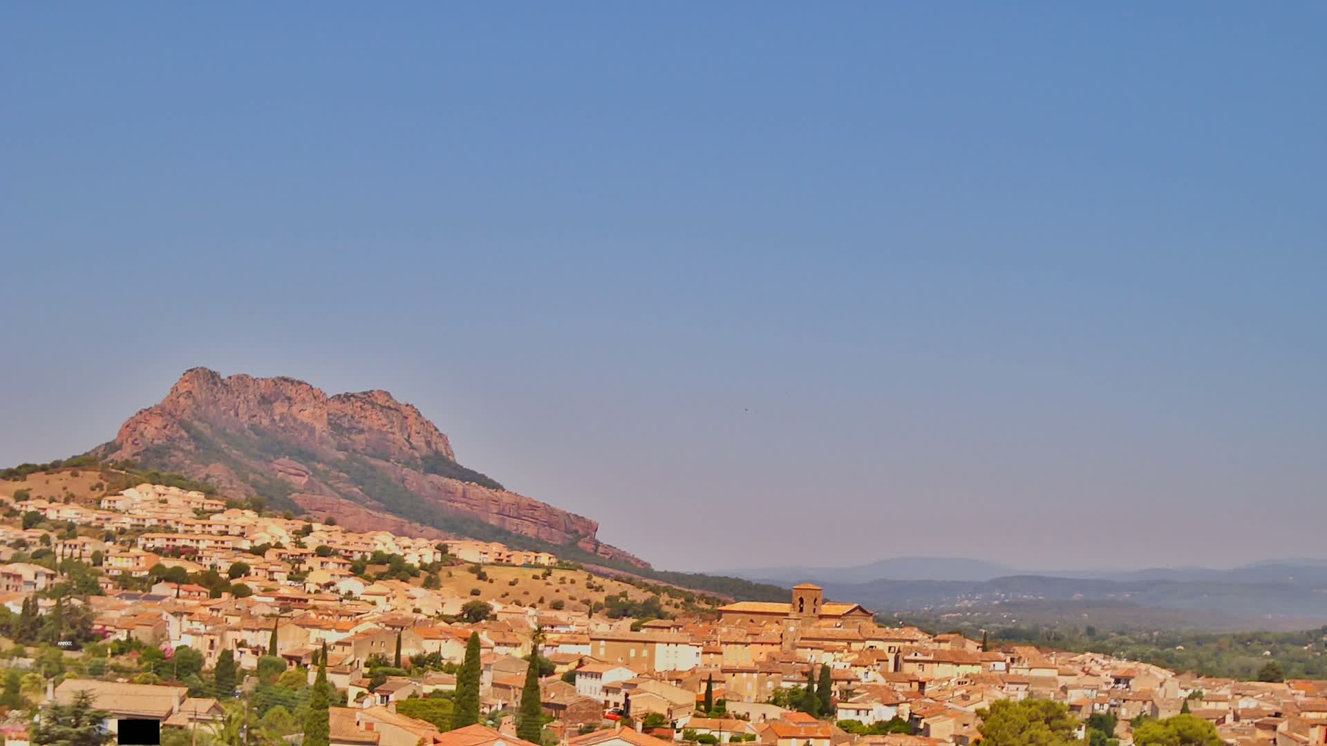 A town of tan and brown buildings sprawls across a hillside beneath a large, reddish-brown mountain under a clear, pale blue sky.